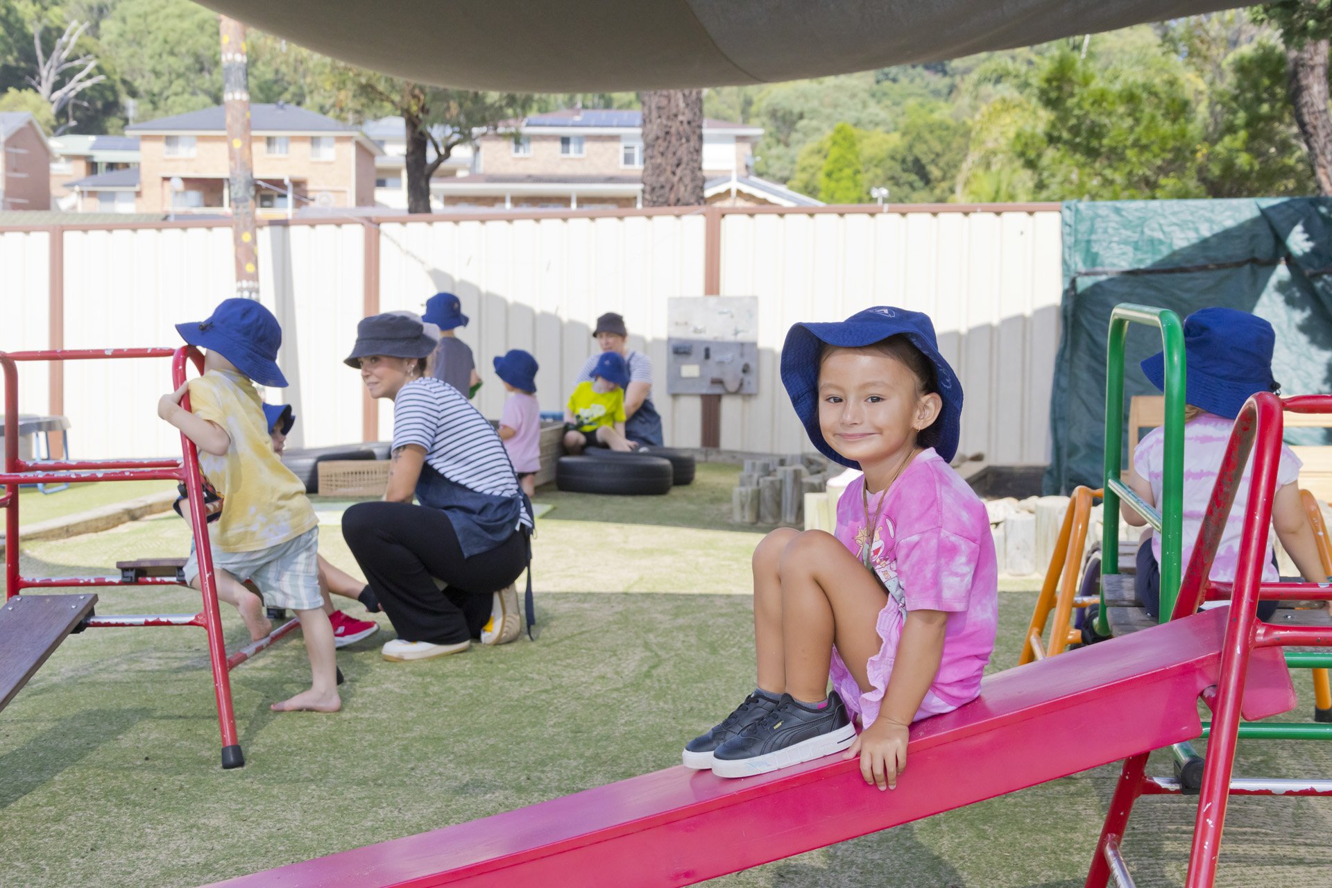 Children and adults playing on playground equipment outdoors, with a girl in a pink tie-dye shirt and blue hat sitting on a pink slide, smiling at the camera.
