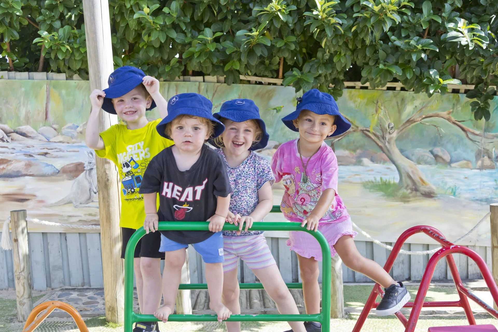 Four children in colorful clothes and blue hats playing on a playground with a painted mural of a river scene in the background.