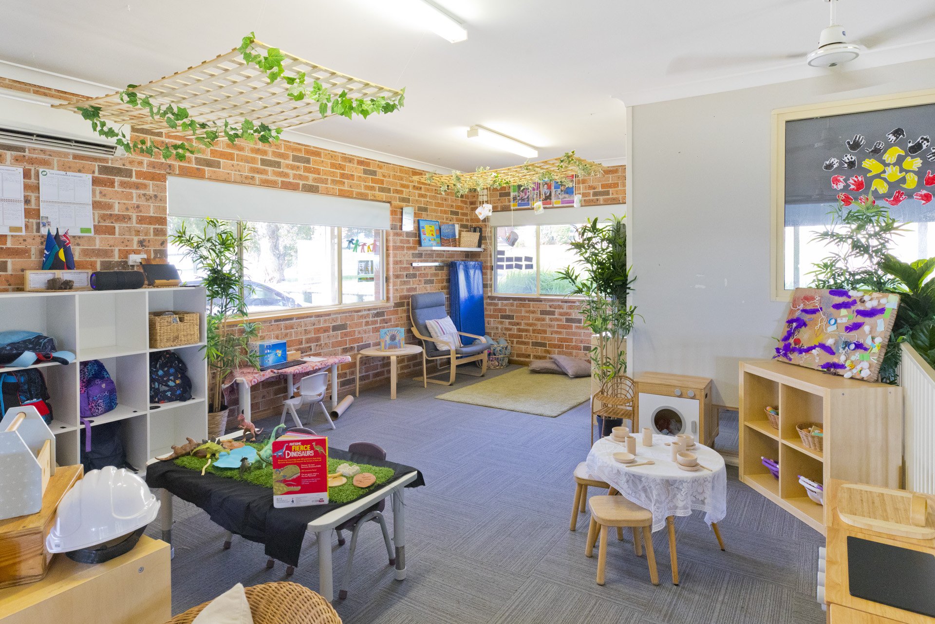 Children's playroom with toys, furniture, and decorations, featuring brick walls, windows, plants, and colorful artwork display.
