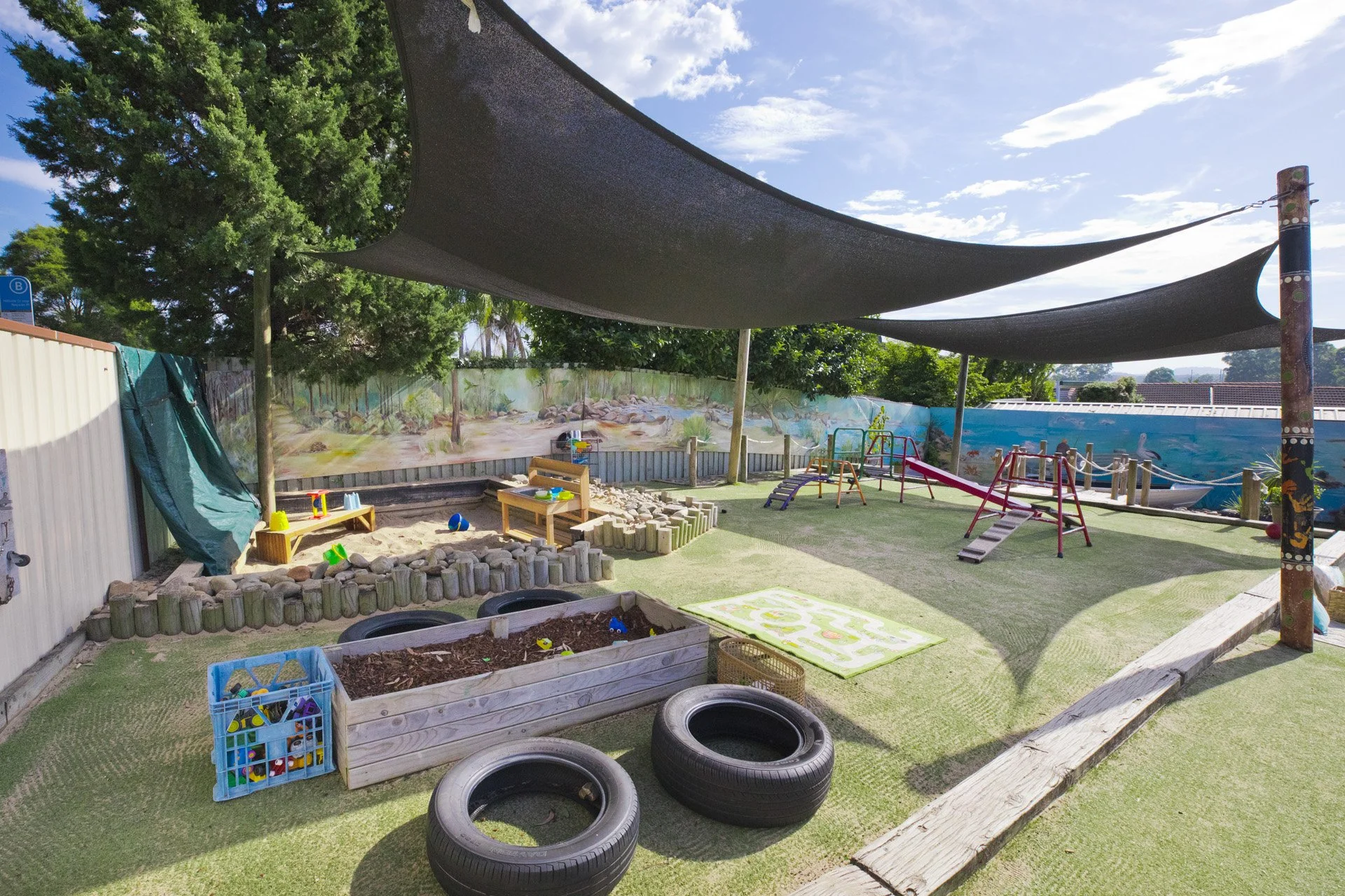 Children's outdoor play area with shade sails, sandbox, wooden logs, tires, climbing frames, and a mural wall in the background.