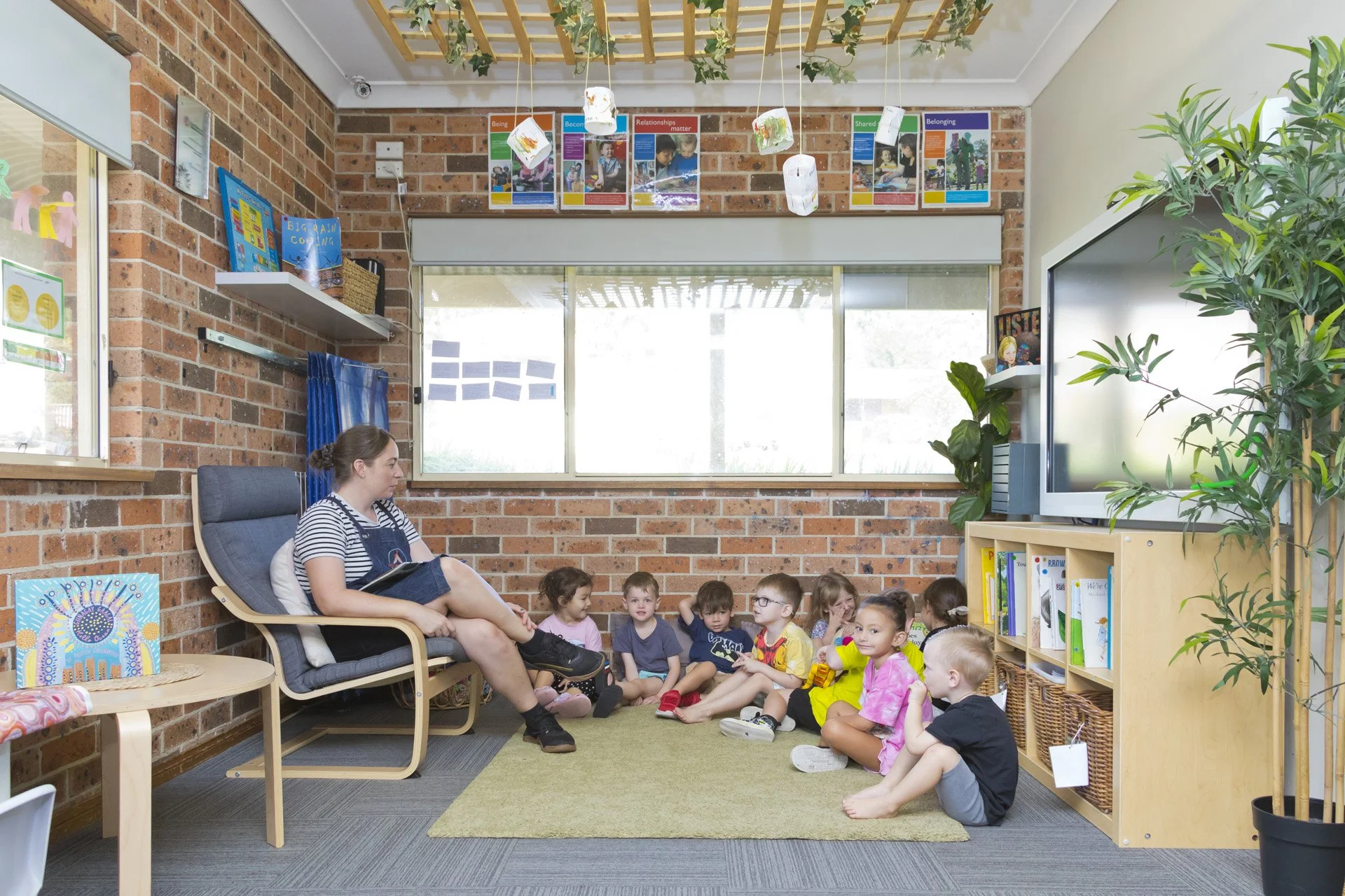 A teacher reading a story to young children in a classroom with brick walls, colorful artwork, and a television on a wooden bookshelf.