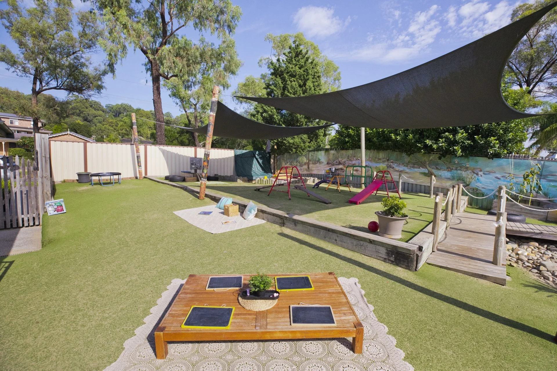 A backyard with artificial grass, outdoor play equipment including slides and swings, shaded by black shade sails, with potted plants, decorative rocks, and a small wooden bridge over a creek, under a sunny sky with trees in the background.
