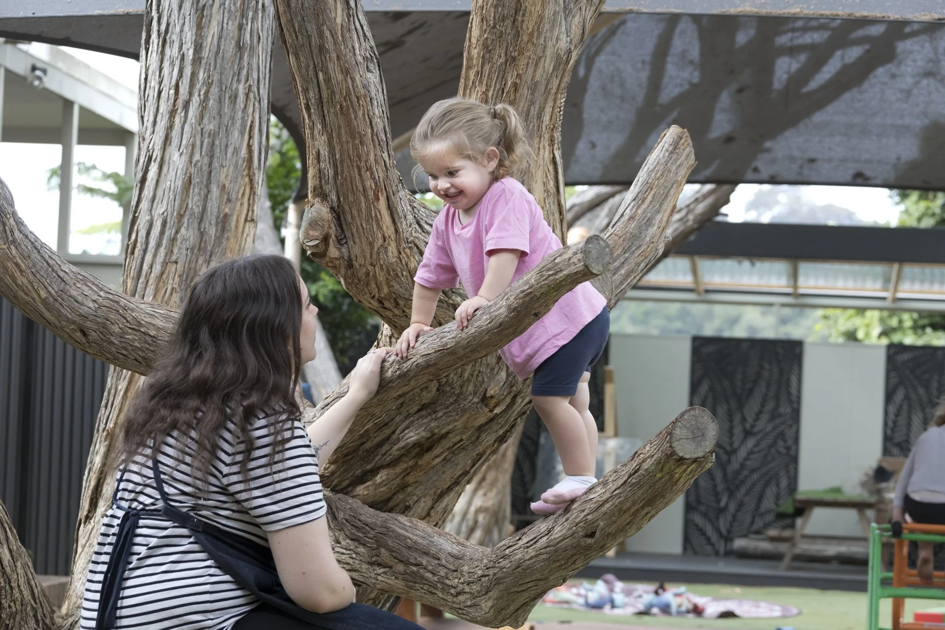 A young girl climbing a large tree with the assistance of an adult woman.