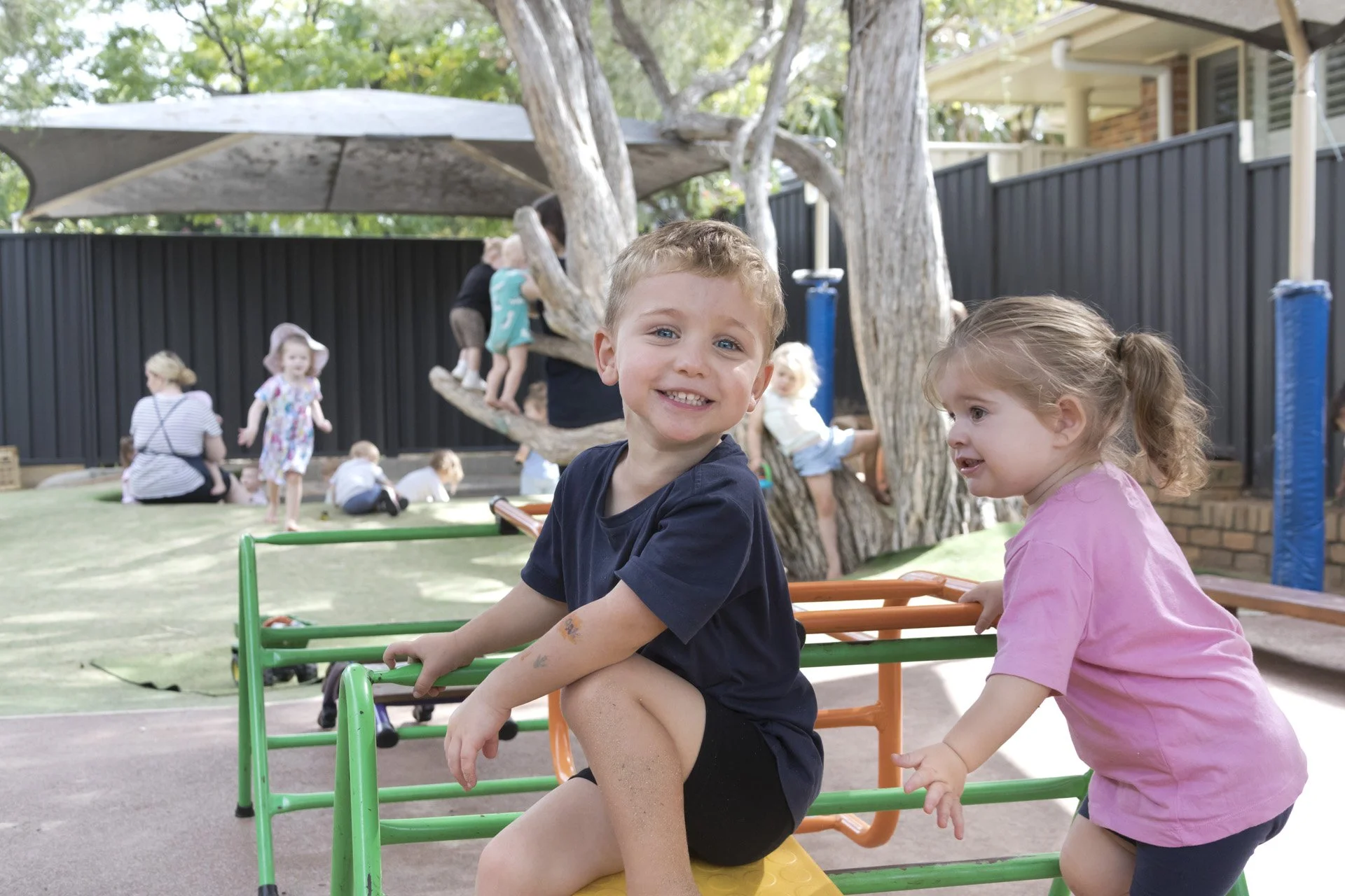 Kids playing on playground equipment, including a boy smiling at the camera and a girl pushing him, with other children and adults playing in the background.