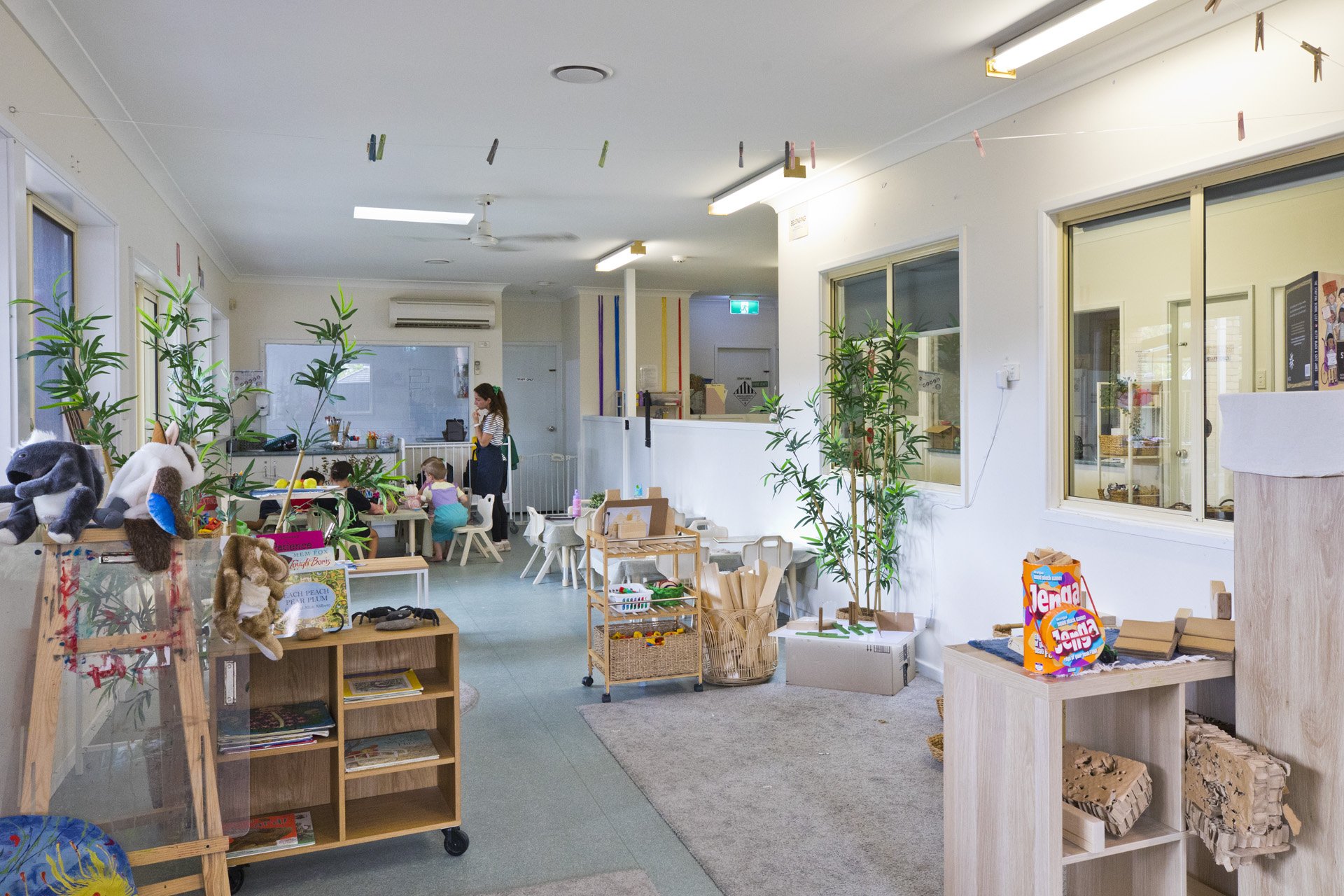 Children playing and a caregiver in a brightly lit indoor playroom decorated with plants, toys, and child-sized furniture.
