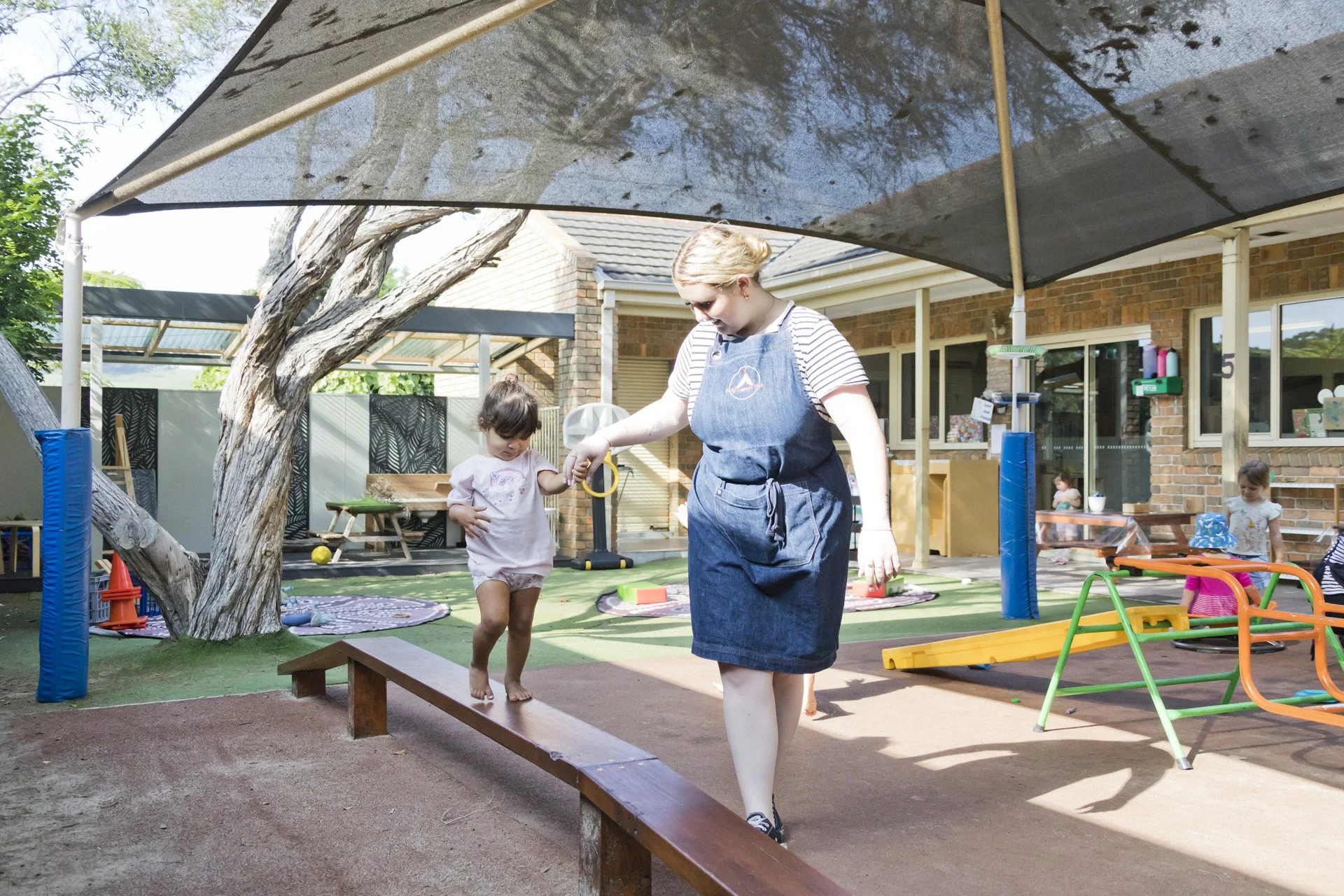 A caregiver helping a young girl walk on a balance beam in an outdoor play area with children playing in the background, a tree, and a brick house.