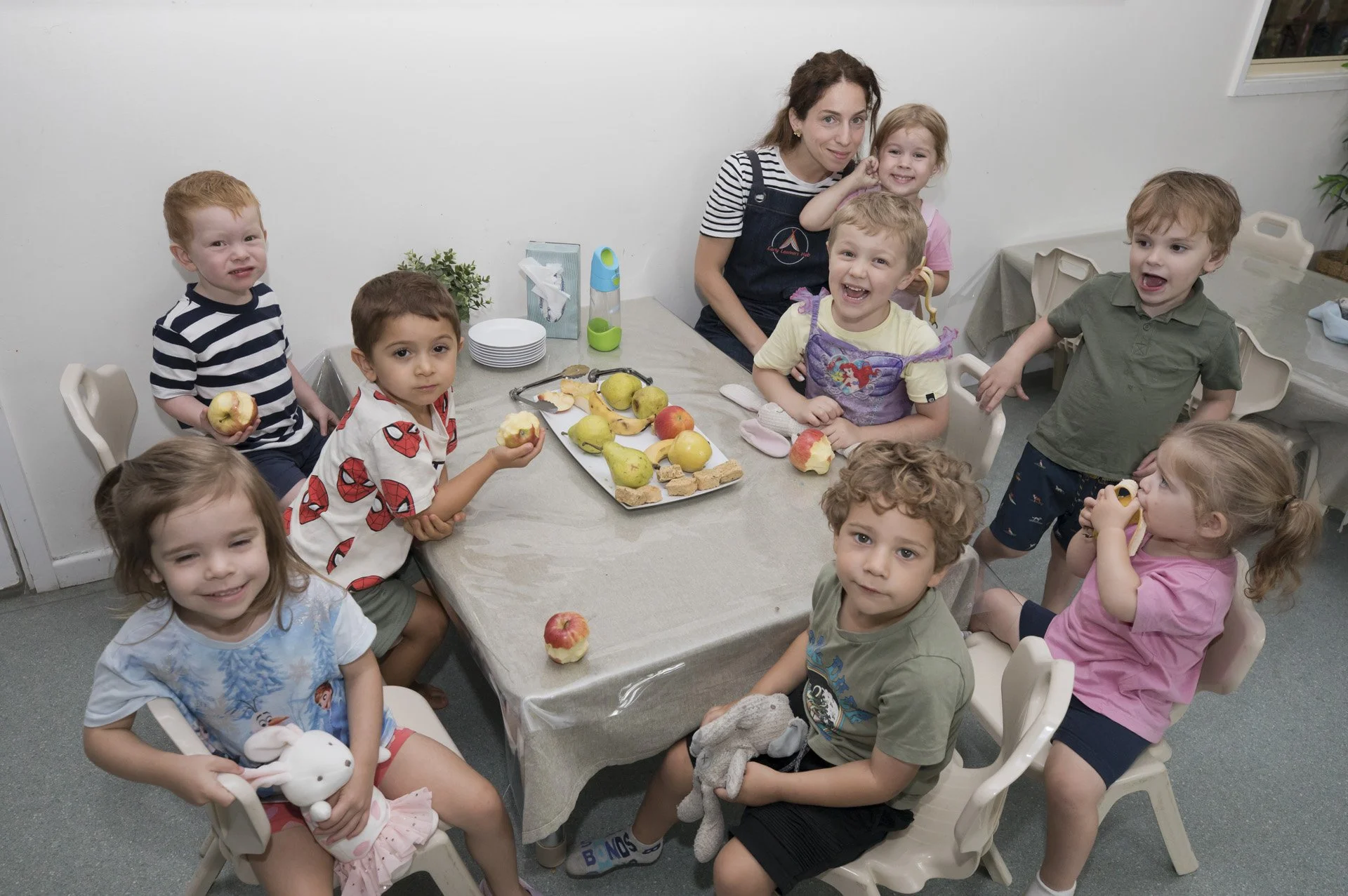 A group of children and an adult woman gathered around a table with apples, snacks, and a few toys, indoors.