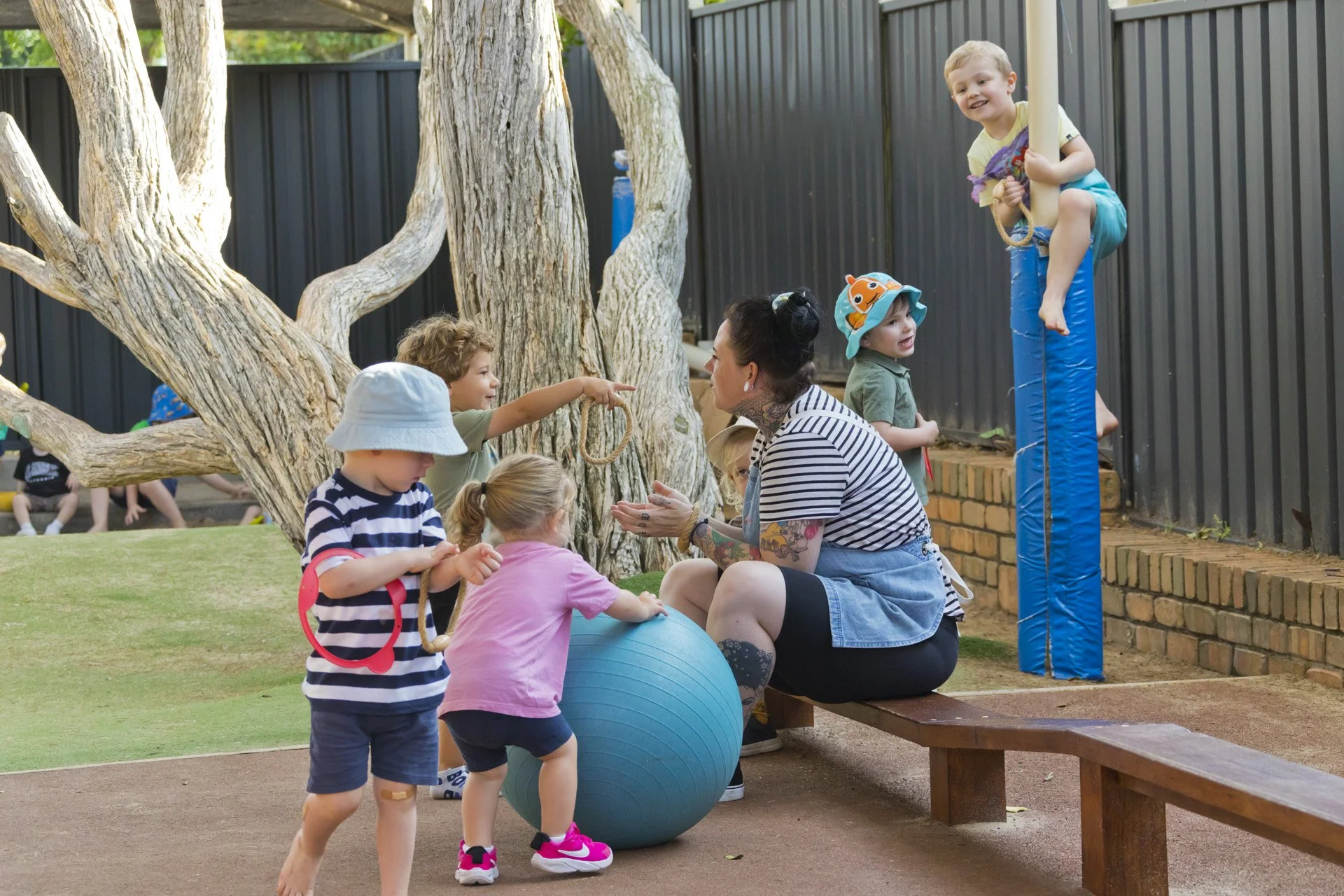 Children playing outside at a park, talking to a woman sitting on a bench, with one child climbing on a pole and another sitting on a blue ball near a tree.