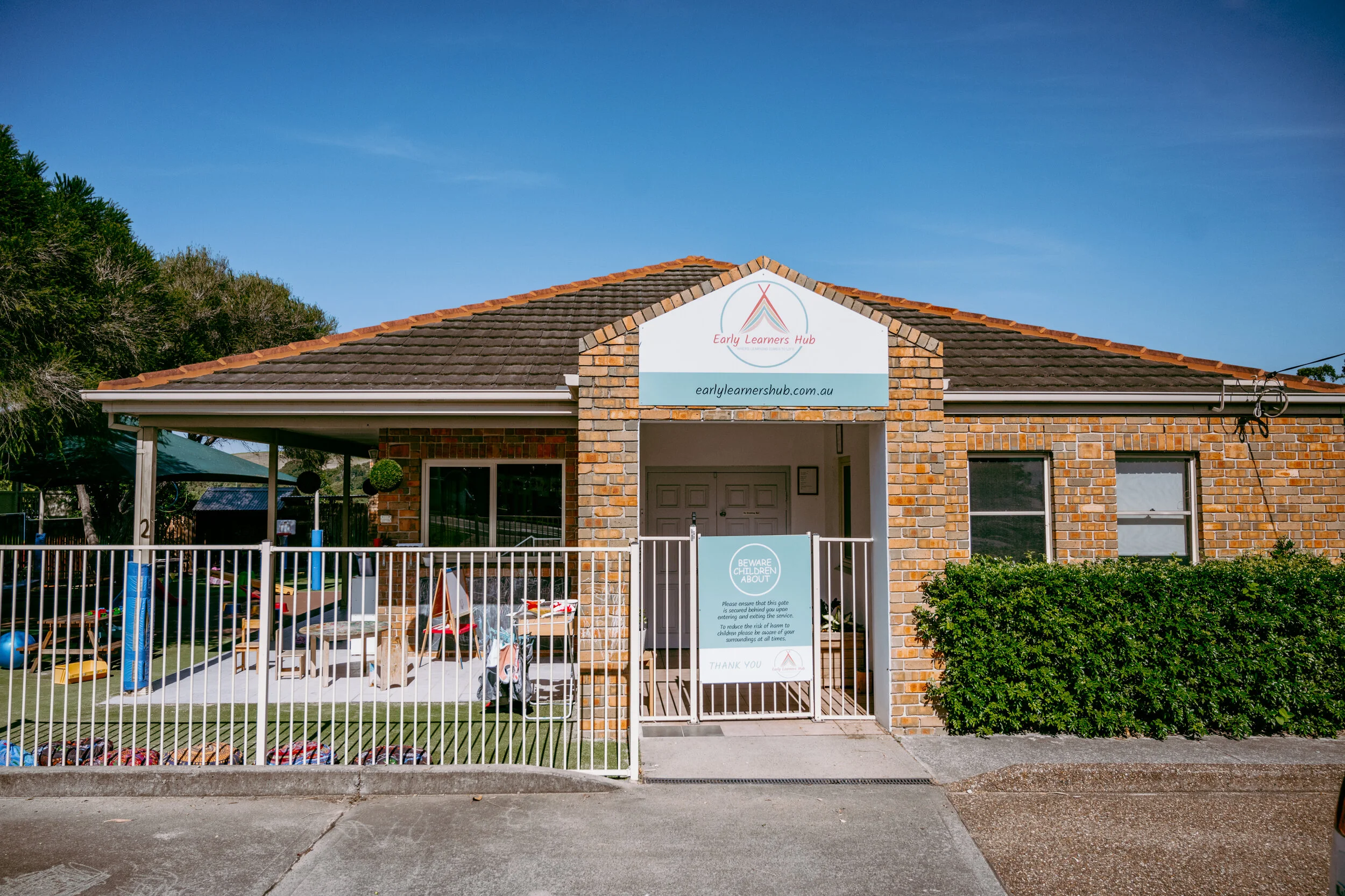 Brick building with a sign for Early Learners Hub, a gated entrance, and outdoor play area with toys and equipment under a clear blue sky.