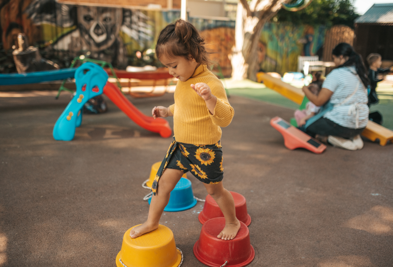 A young girl in a yellow sweater and sunflower-patterned shorts balancing on stepping stones in a playground.