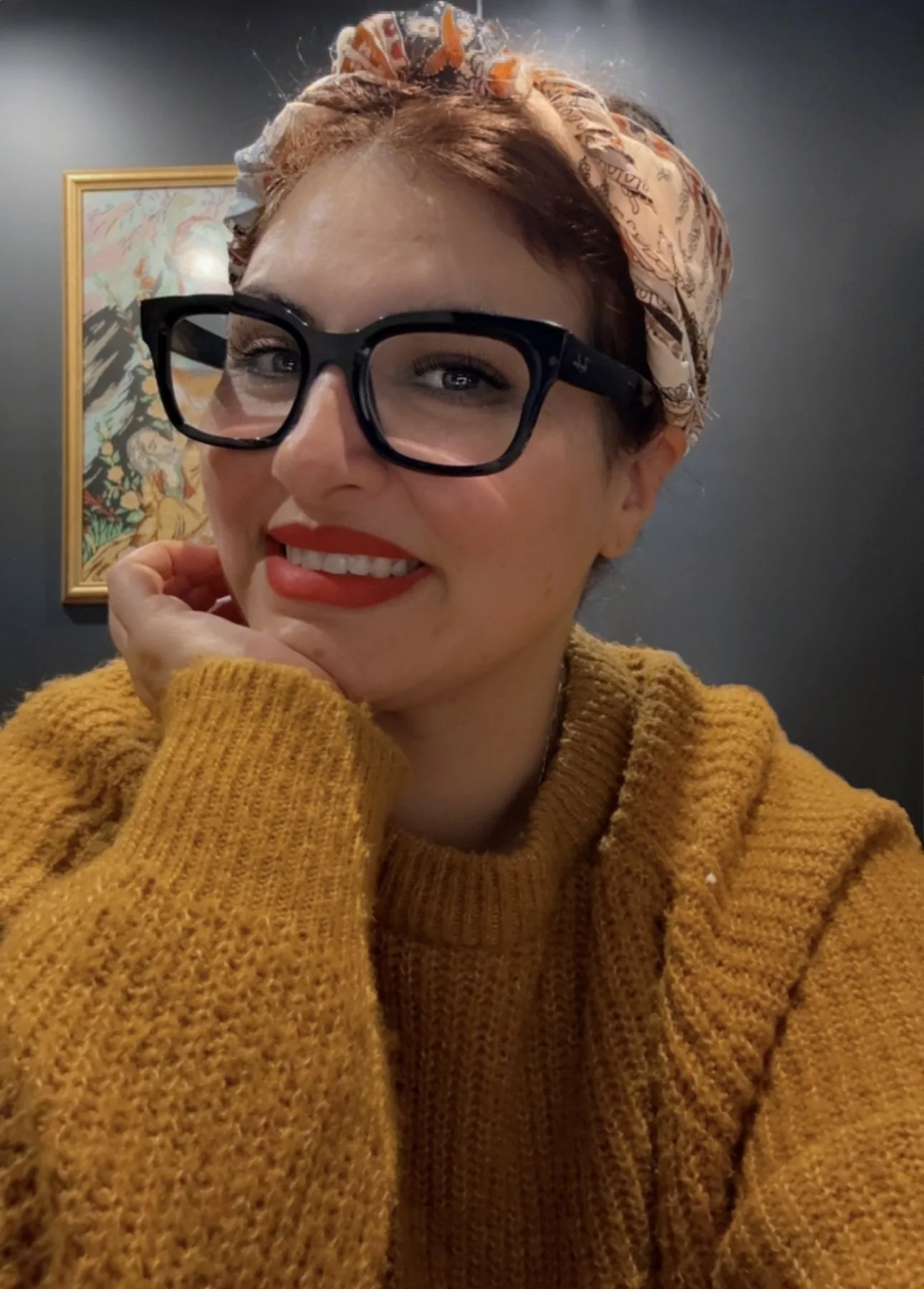 A young woman with red hair wearing a patterned headscarf, large black glasses, red lipstick, and a mustard-colored sweater, smiling with her hand resting on her chin, in front of a dark wall with a framed abstract artwork.