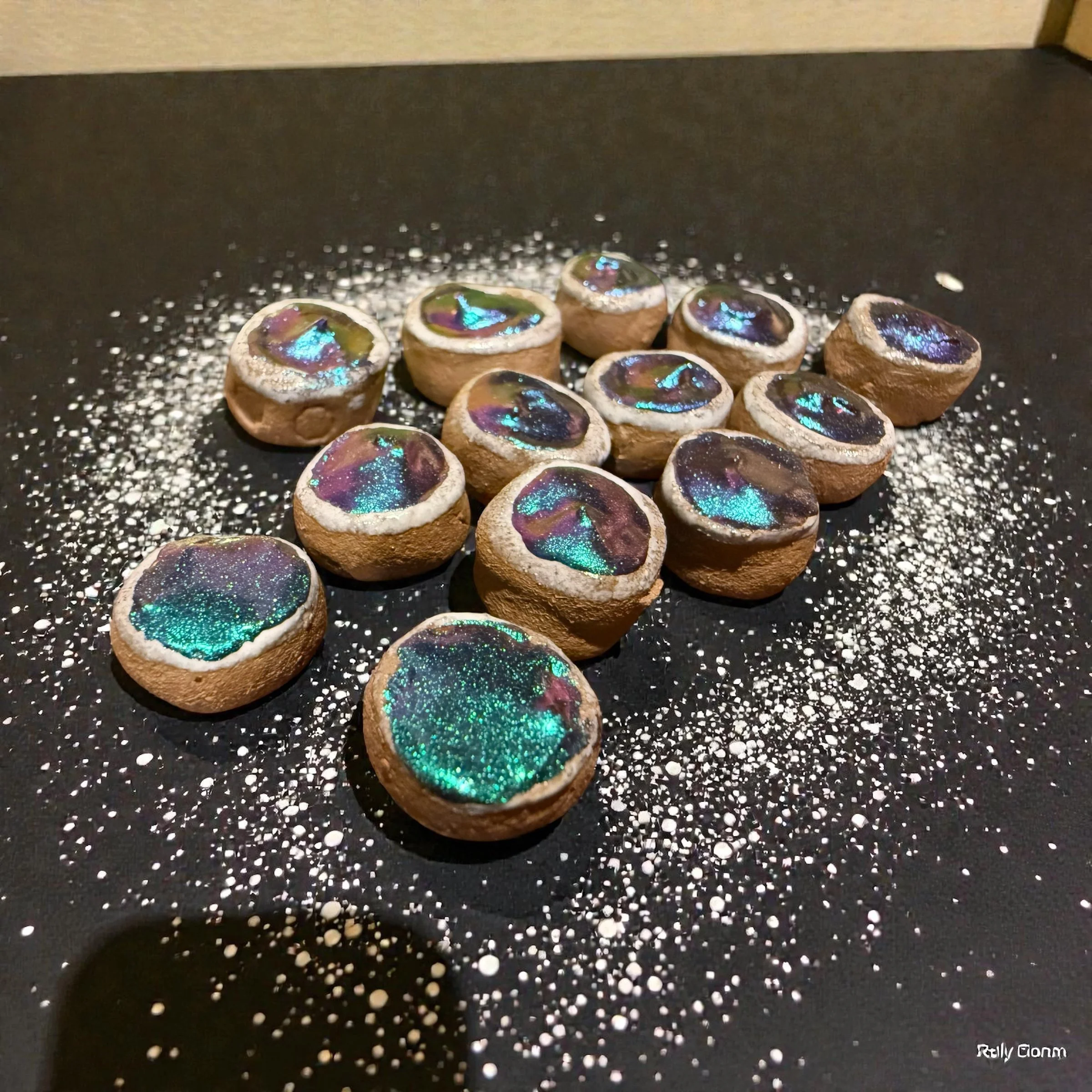 Small round cookies topped with iridescent, galaxy-like edible decorations, arranged on a black surface scattered with white powdered sugar.