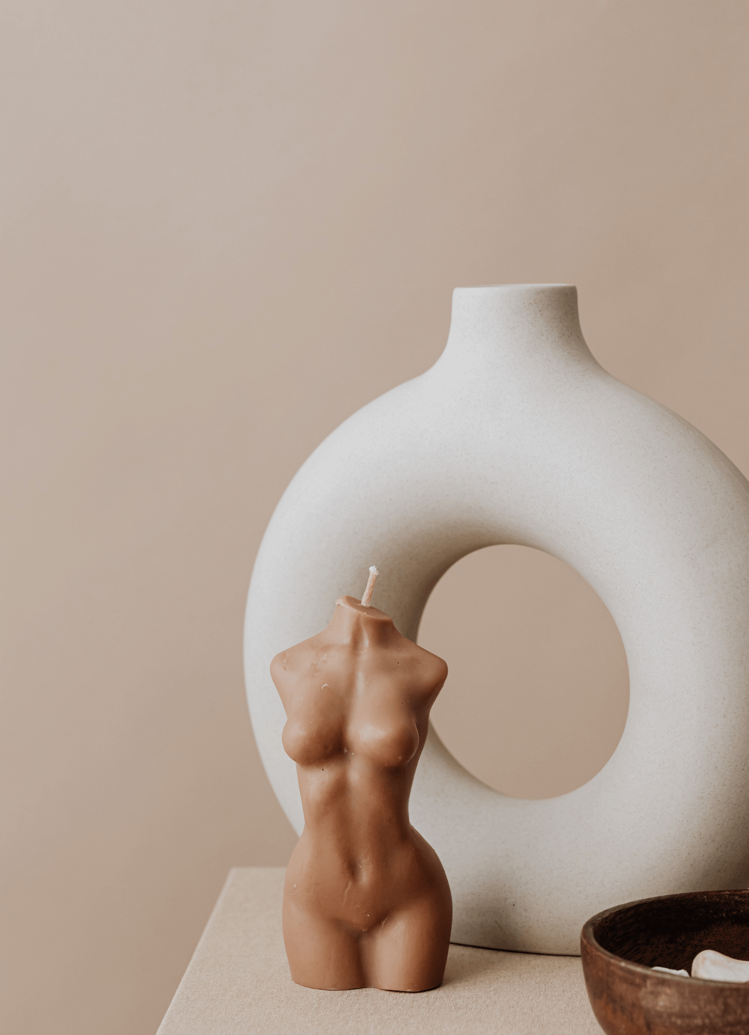 Decorative scene with a large white textured vase with a hole in the center, a body-shaped candle, and a wooden bowl on a beige surface against a beige background.