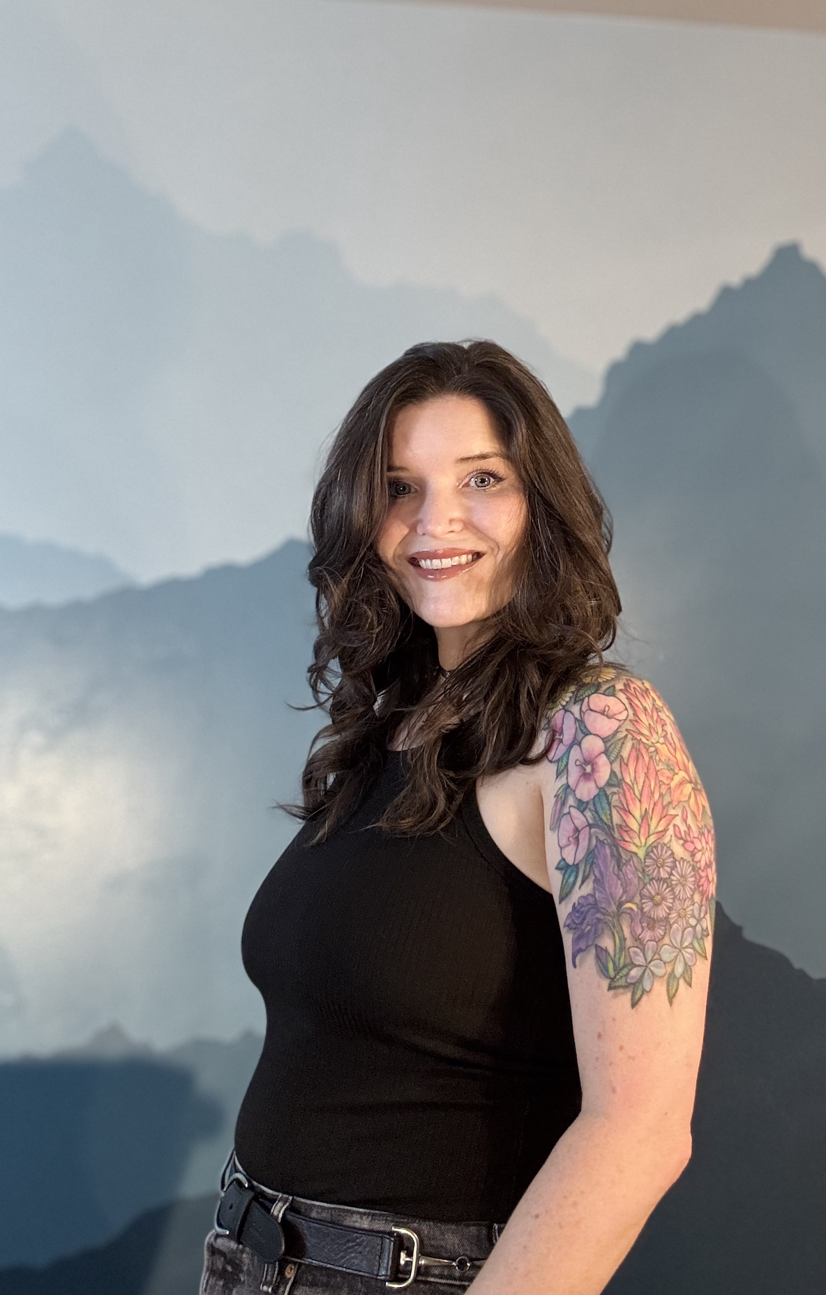 A woman with dark brown hair, smiling, wearing a black sleeveless top and jeans, standing in front of a painted mountain landscape background, showing a colorful floral tattoo on her upper arm.