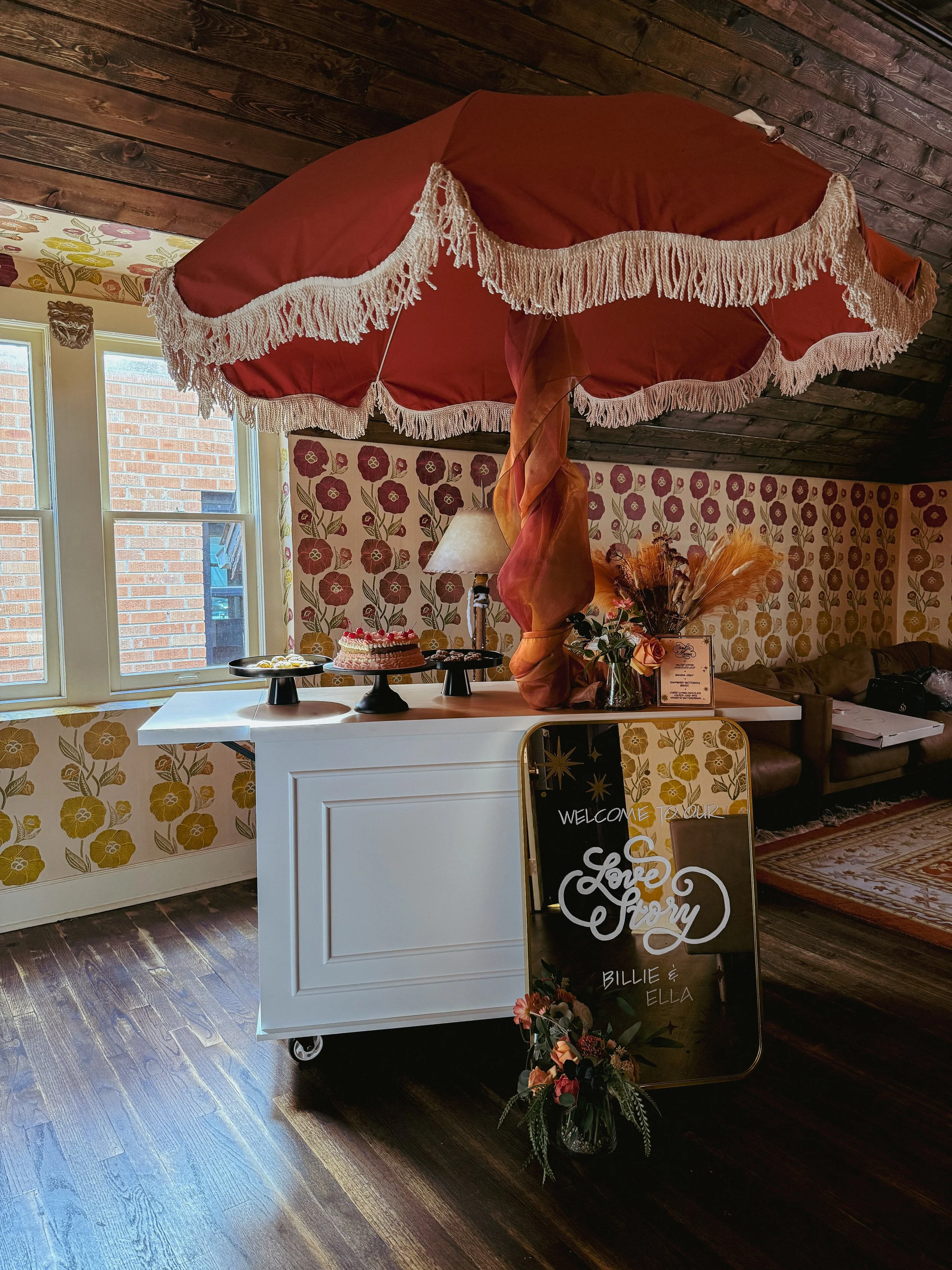 A decorated interior with a large red parasol with white fringes, a white counter with desserts, and a welcome sign for Billie and Ella with flowers.