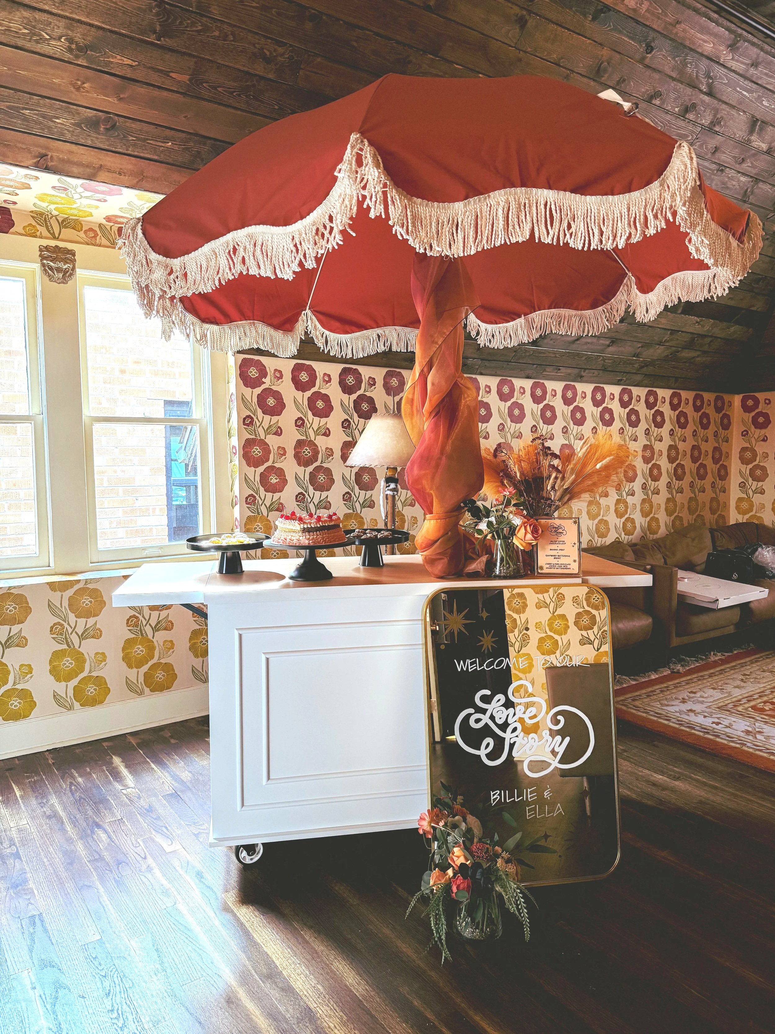 Decorative welcome sign at a wedding reception featuring a mirror with 'Love Story Billie & Ella' written on it, placed on the floor beside a floral arrangement in a vase. The setup is under a large vintage-style red umbrella with a fringed edge, situated on a white cart. The background includes floral-patterned wallpaper, a window, a lamp, and a corner of a sofa with cushions.