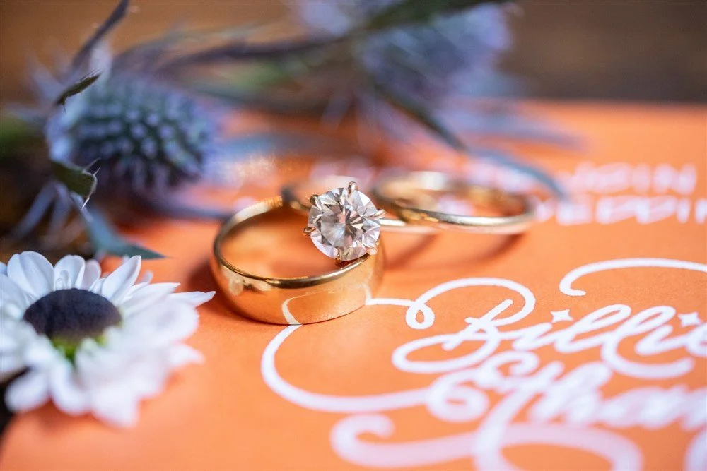 A close-up of wedding rings, one with a large diamond, placed on an orange card with white cursive text, surrounded by a white daisy flower and green greenery.