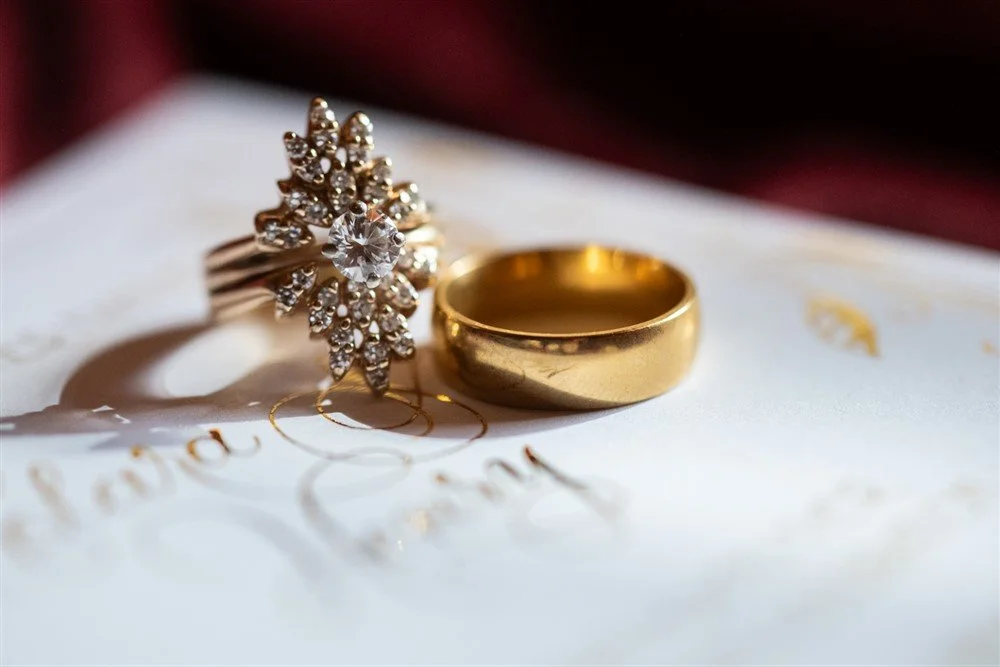 Close-up of two wedding rings, a gold band and a diamond engagement ring with a floral design and center stone, resting on an invitation card.