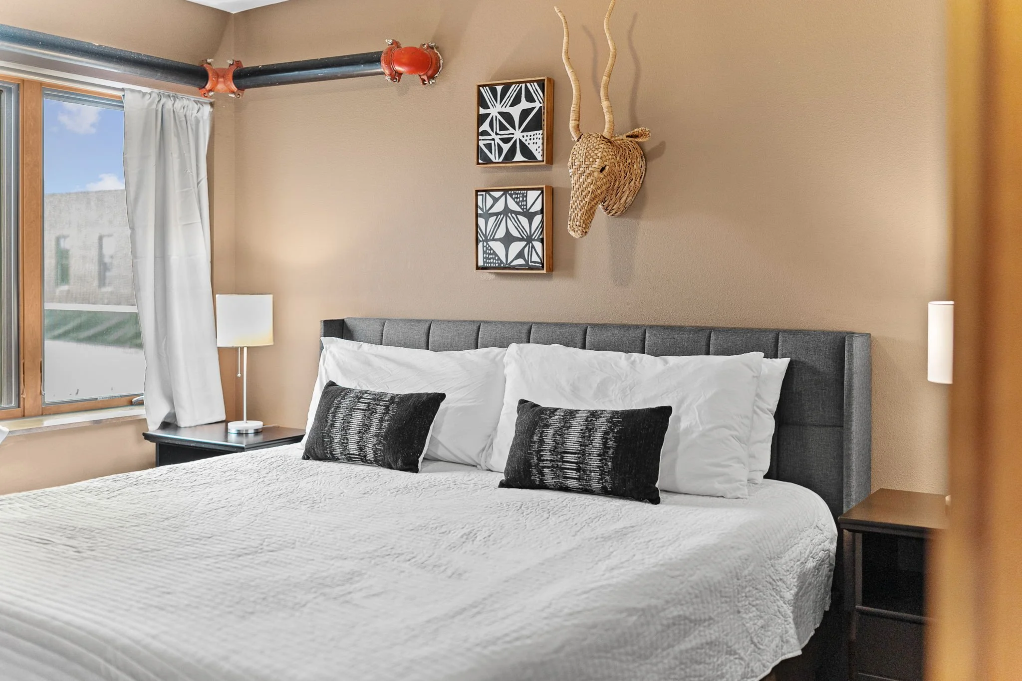 A bedroom with a large bed with white bedding and black textured pillows, a gray upholstered headboard, a nightstand with a white lamp, beige walls, wall art, a window with white curtains, and visible industrial piping on the ceiling.
