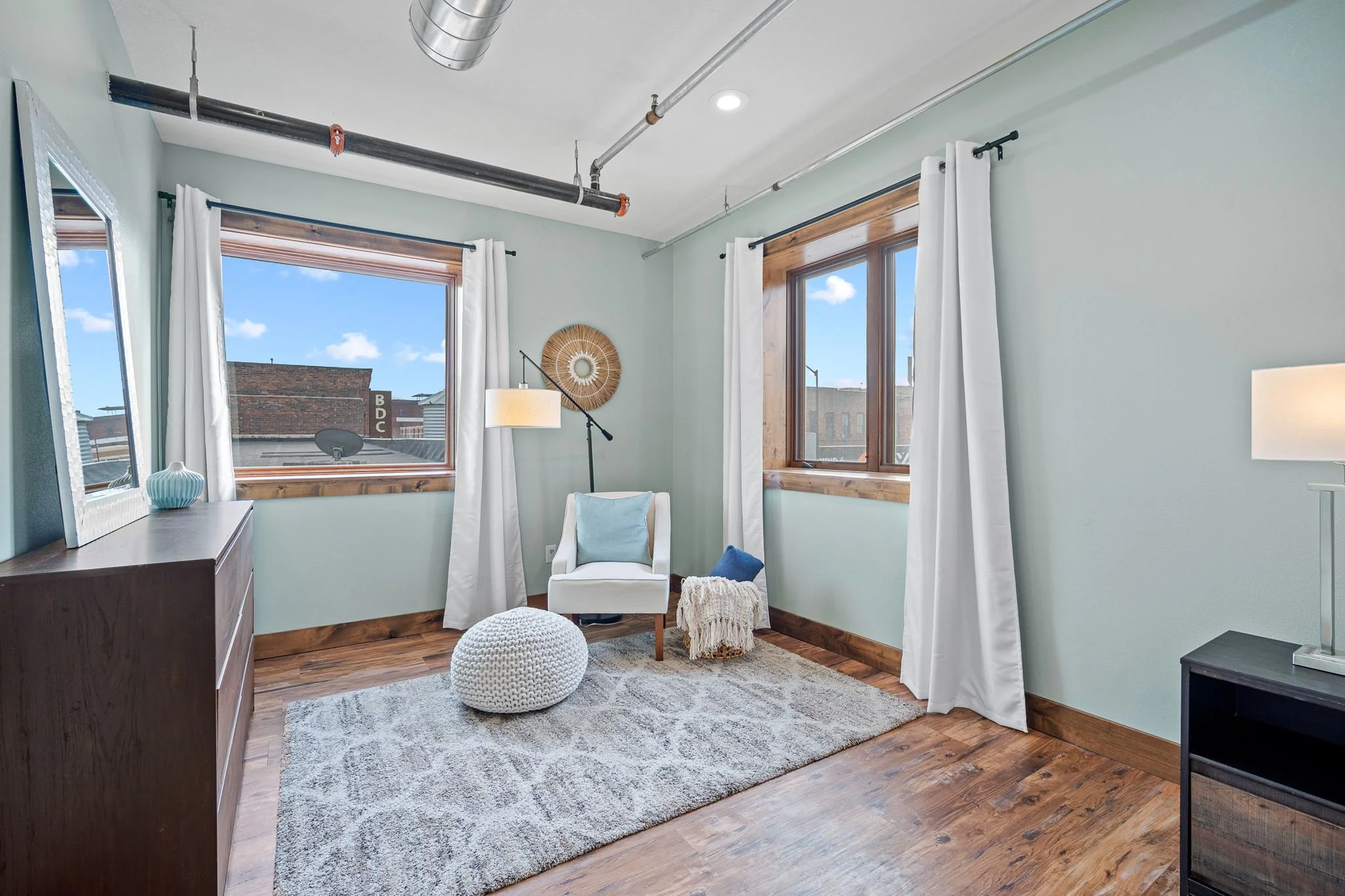 A cozy living room with two large windows with white curtains, a white armchair with a light blue pillow, a beige knitted pouf, a wooden dresser, and decorative items including a mirror and a blue vase. The ceiling features exposed black pipes and ductwork, and the room has hardwood floors and a light pastel wall color.