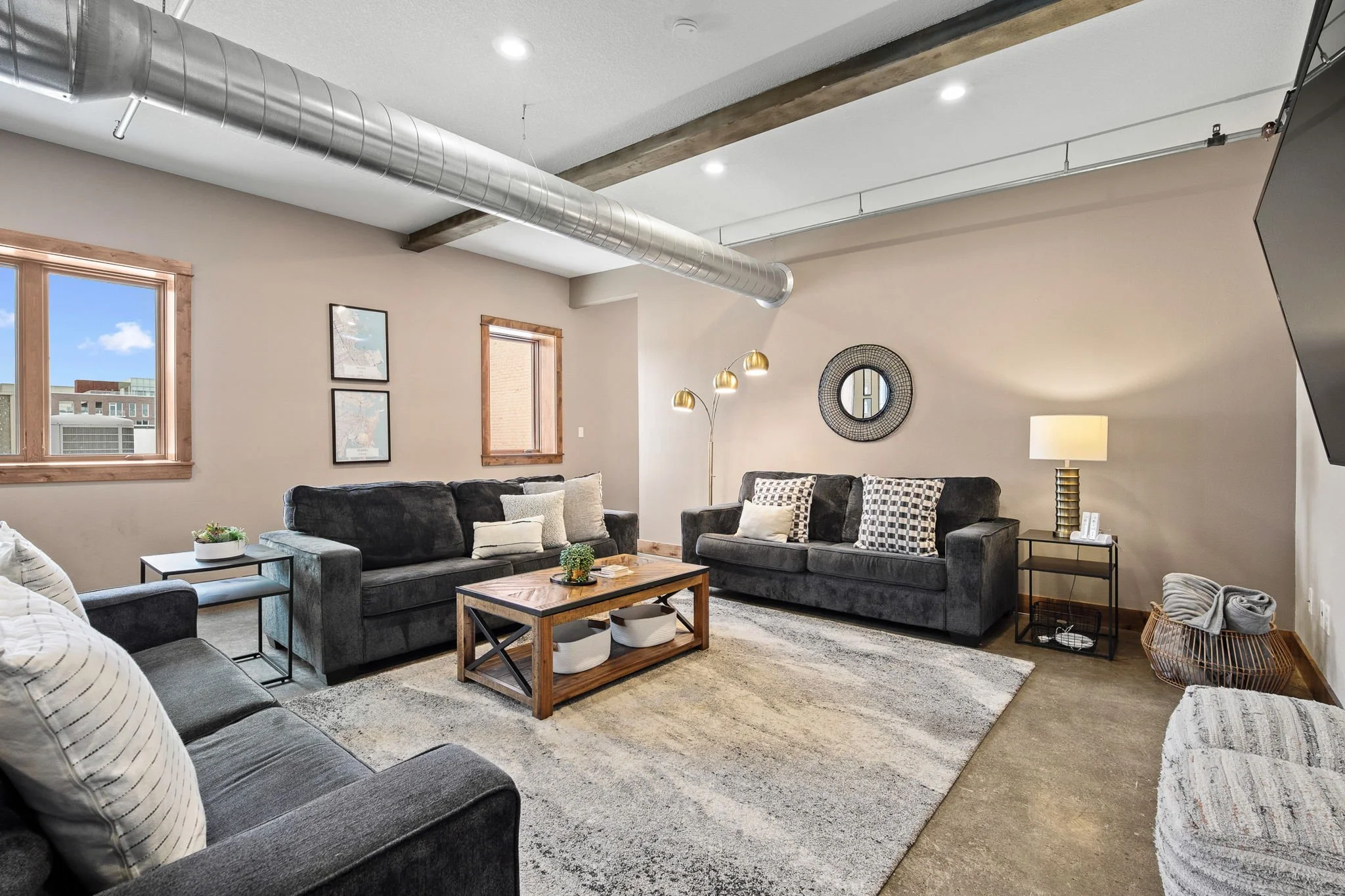 Living room with two gray couches, cushions, a wooden coffee table, a rug, lamps, a decorative mirror, windows, and artwork on the walls.
