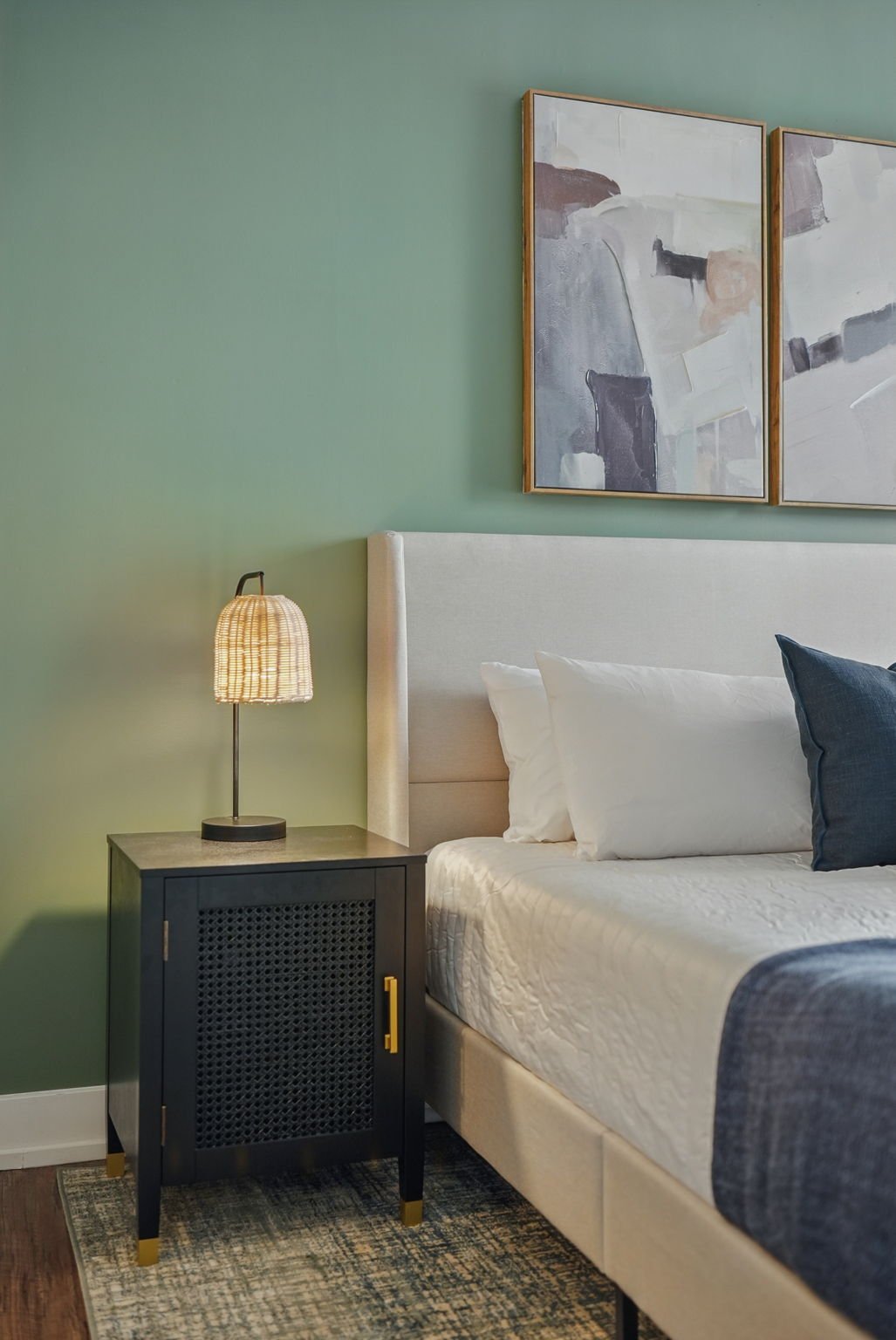 A bedroom with a light green wall, abstract artwork, a beige headboard, and a black bedside table with a wicker table lamp.