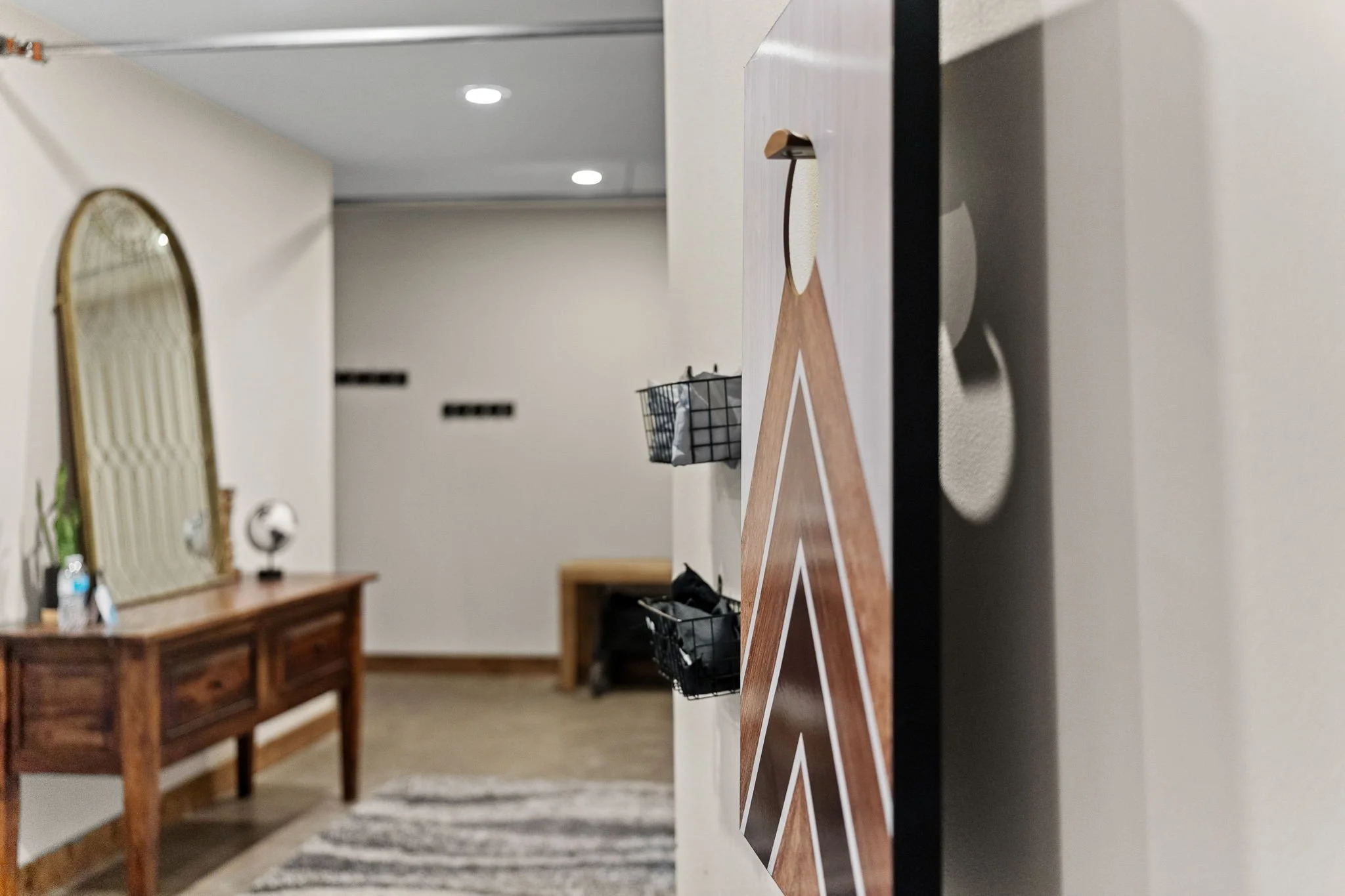 Interior of a home featuring a wooden sideboard with a mirror, a vase, and decorative items. In the background, there are two black wall-mounted baskets, a patterned rug, and a wall with small black rectangular details. The image is taken from a uniq