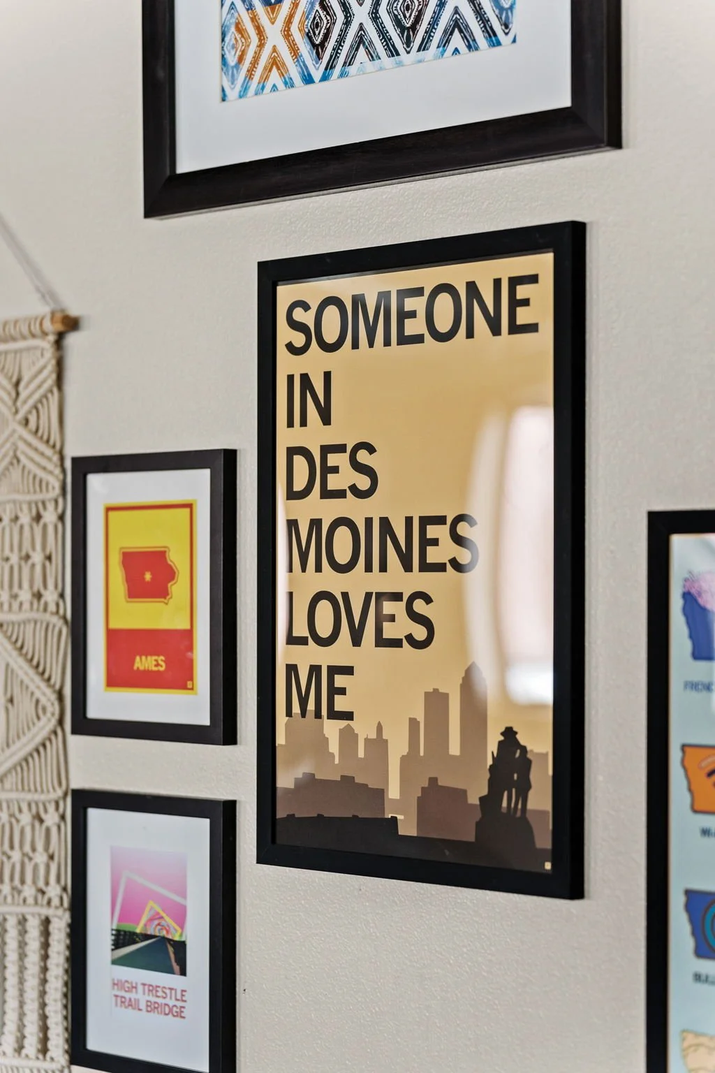 Decorative wall with framed artwork, including a poster that reads "SOMEONE IN DES MOINES LOVES ME," with a silhouette of a city skyline and a statue. Other frames display a stylized map of Arkansas labeled "AMES" and a colorful artwork titled "HIGH 