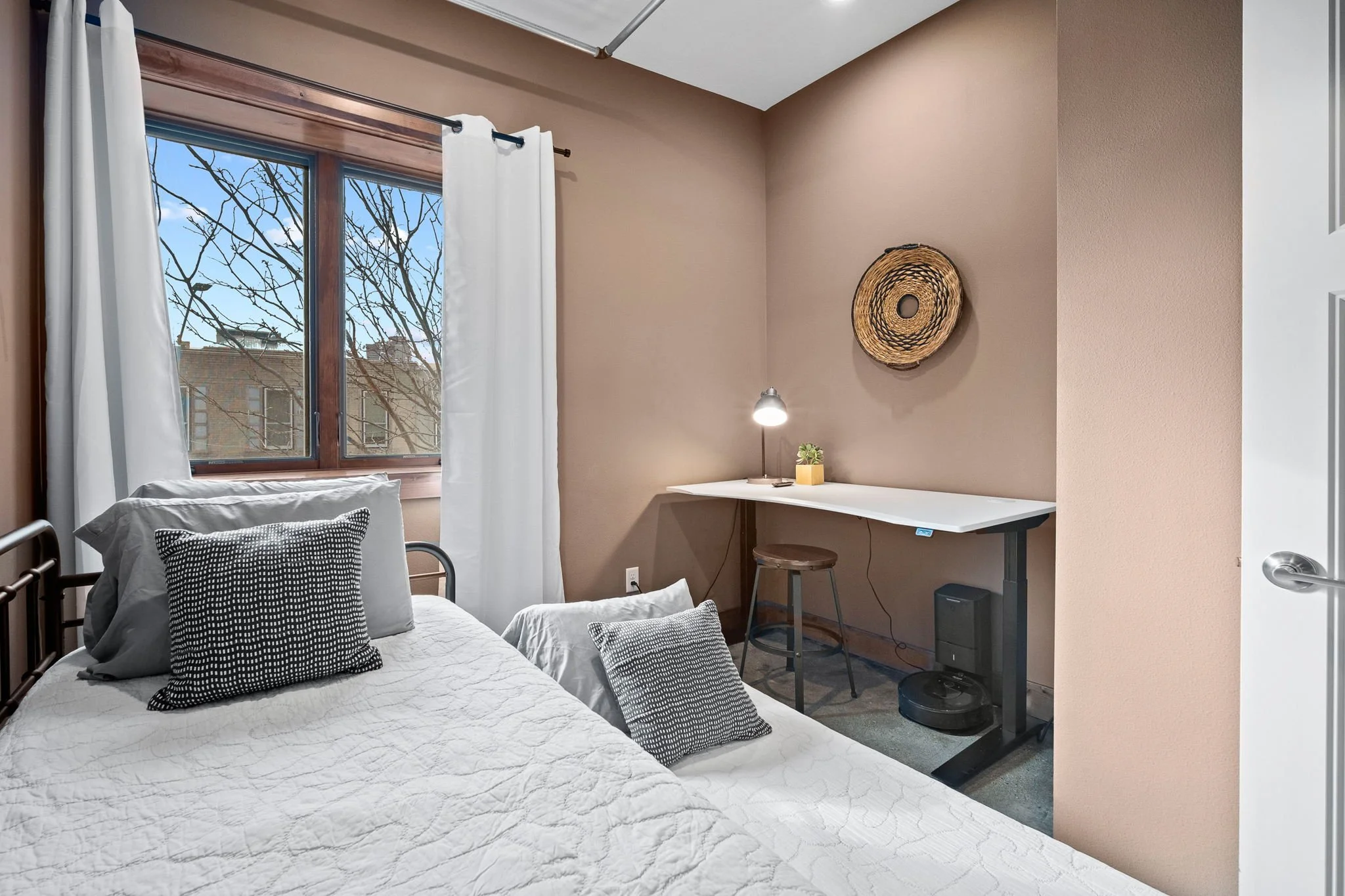 A bedroom with a bed on the left, decorated with black-and-white pillows. There is a window with white curtains and a view of trees and a building outside. A small desk with a lamp, plant, and stool is against the beige wall, which features a round woven wall art piece. Part of a door with a silver handle is visible on the right.