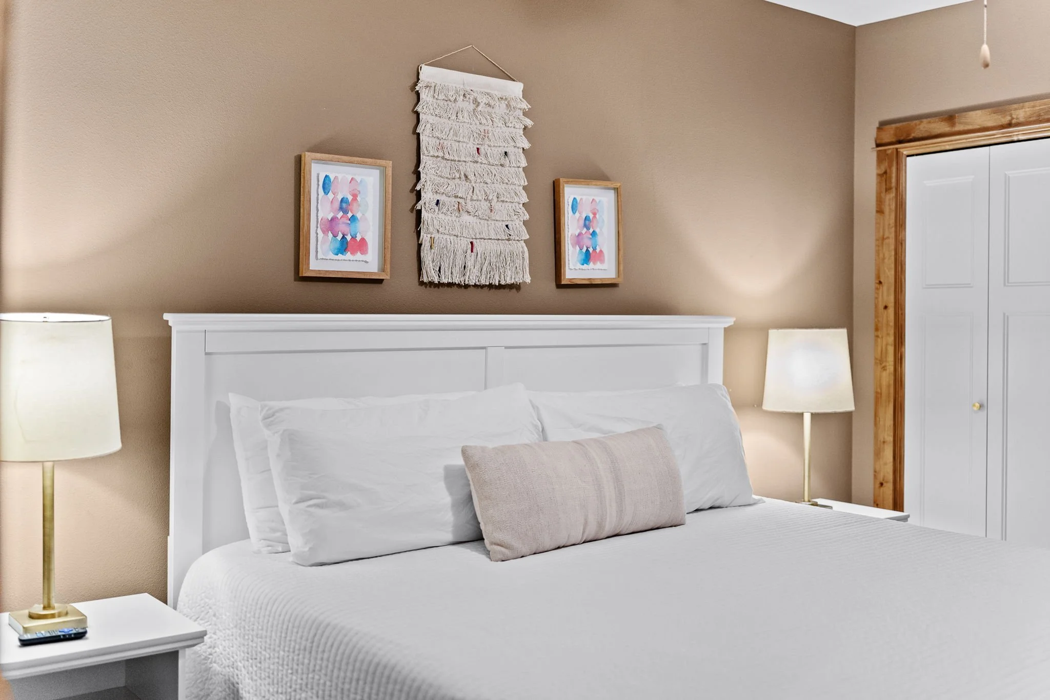 A bedroom with a white bed and pillows, beige walls, two framed watercolor art prints, a woven wall hanging, two table lamps, and a bathroom door with wooden trim.