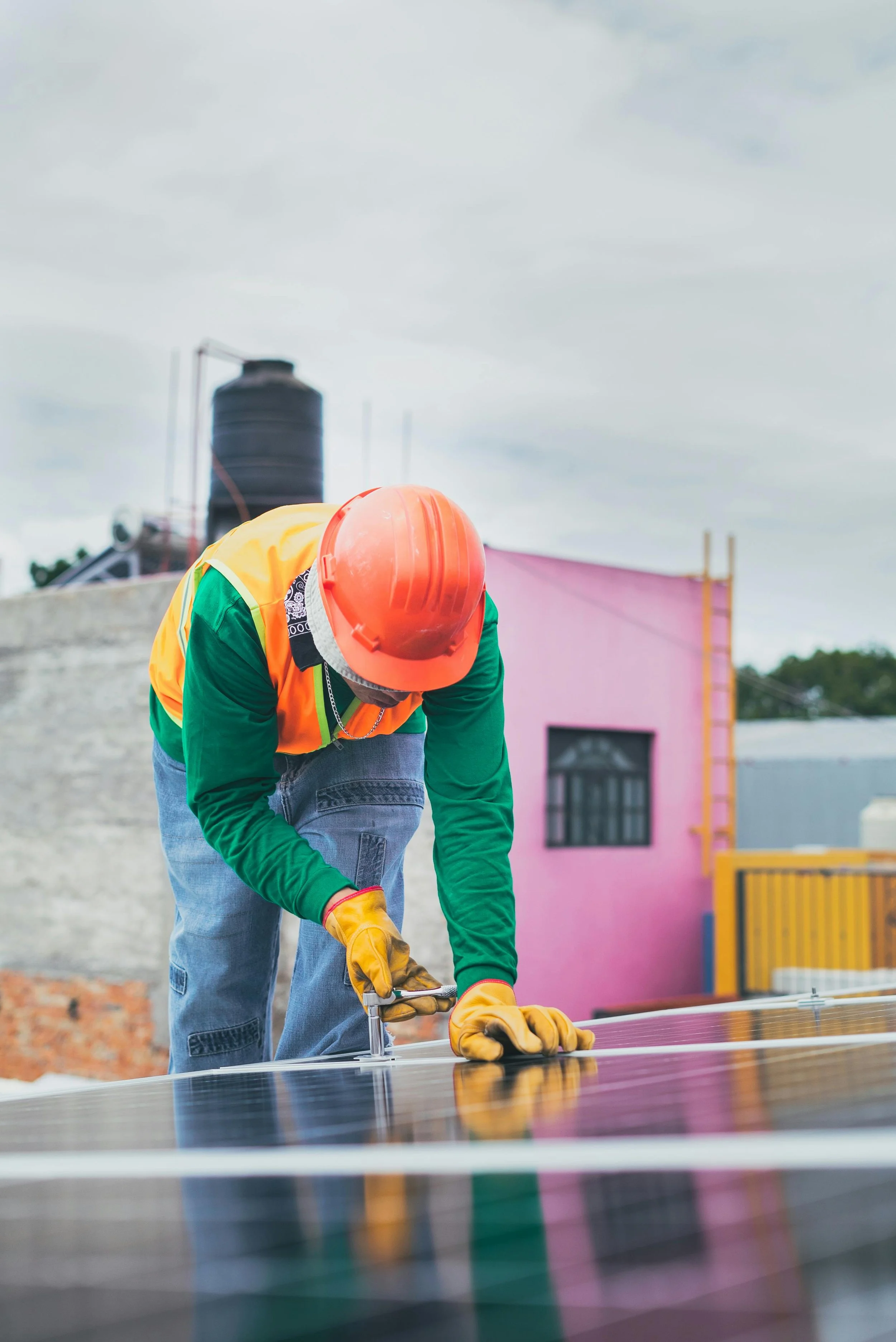 Worker installing solar panels outdoors, wearing an orange hard hat, green jacket with yellow safety vest, and yellow gloves.