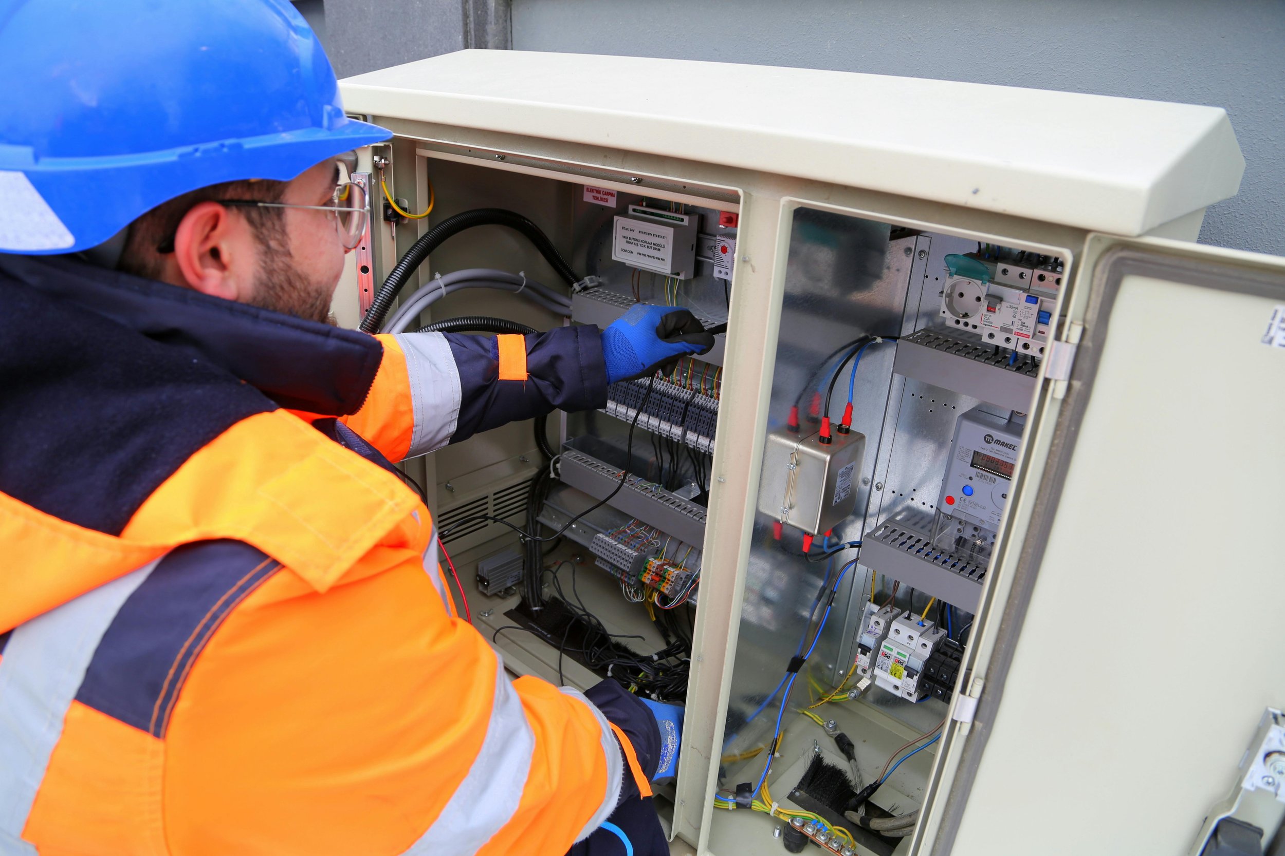 A technician wearing a blue hard hat, safety glasses, gloves, and an orange safety jacket working inside an electrical control cabinet.