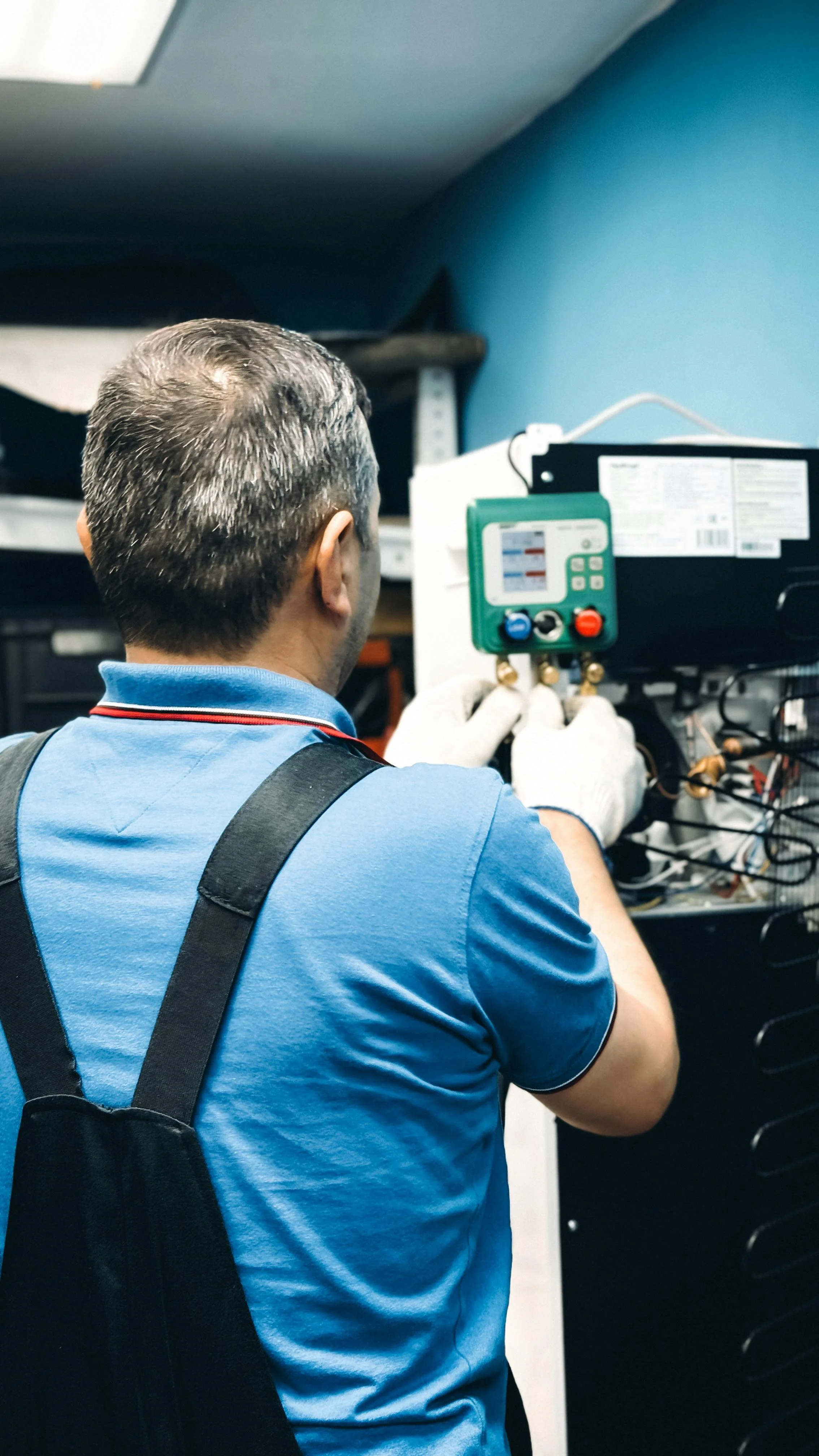 A technician in a blue shirt and white gloves working on an electrical control panel with a testing device.