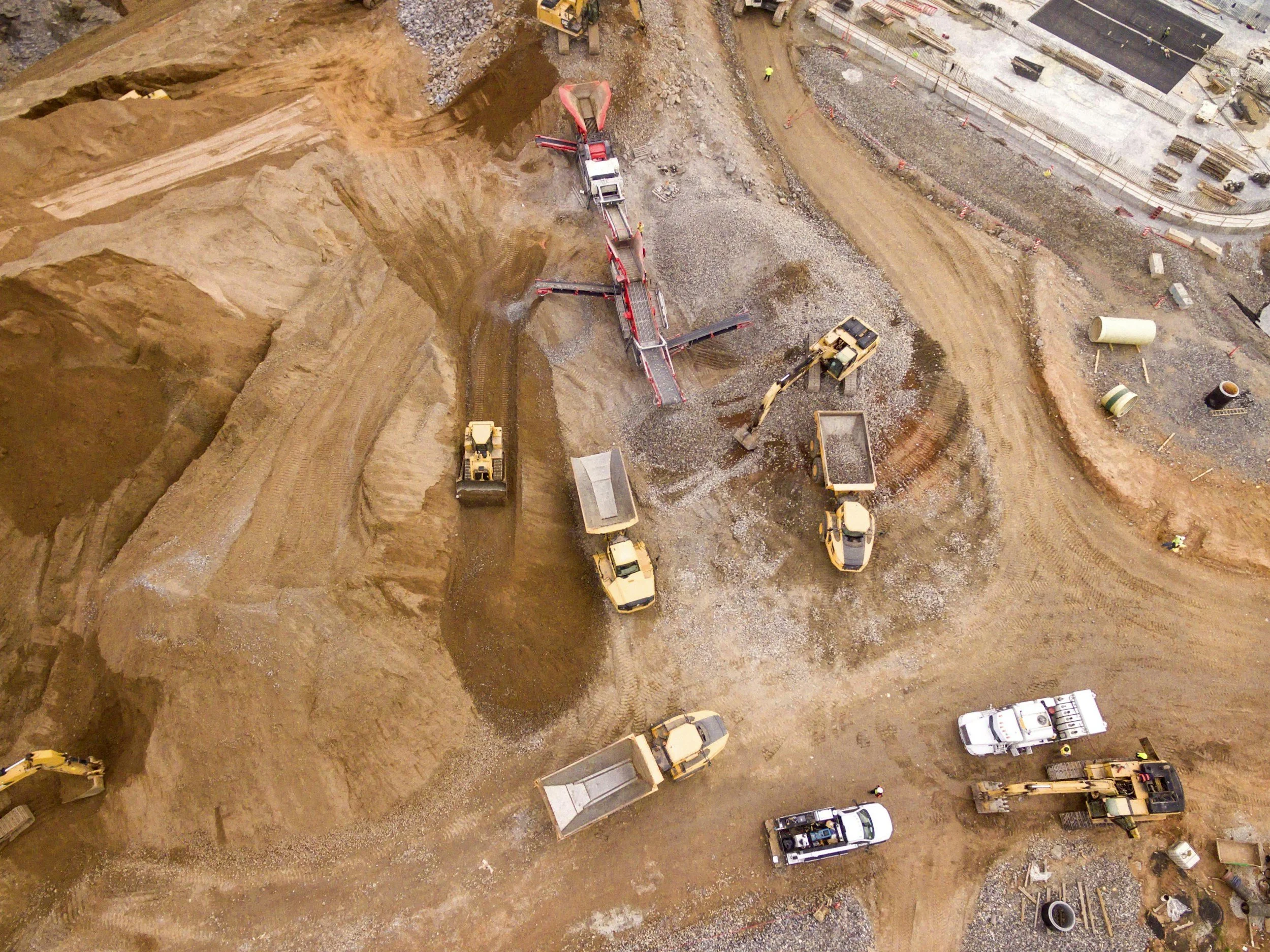 An aerial view of a construction site with multiple bulldozers, excavators, trucks, and construction materials. Large areas of dirt and gravel are being moved and shaped.