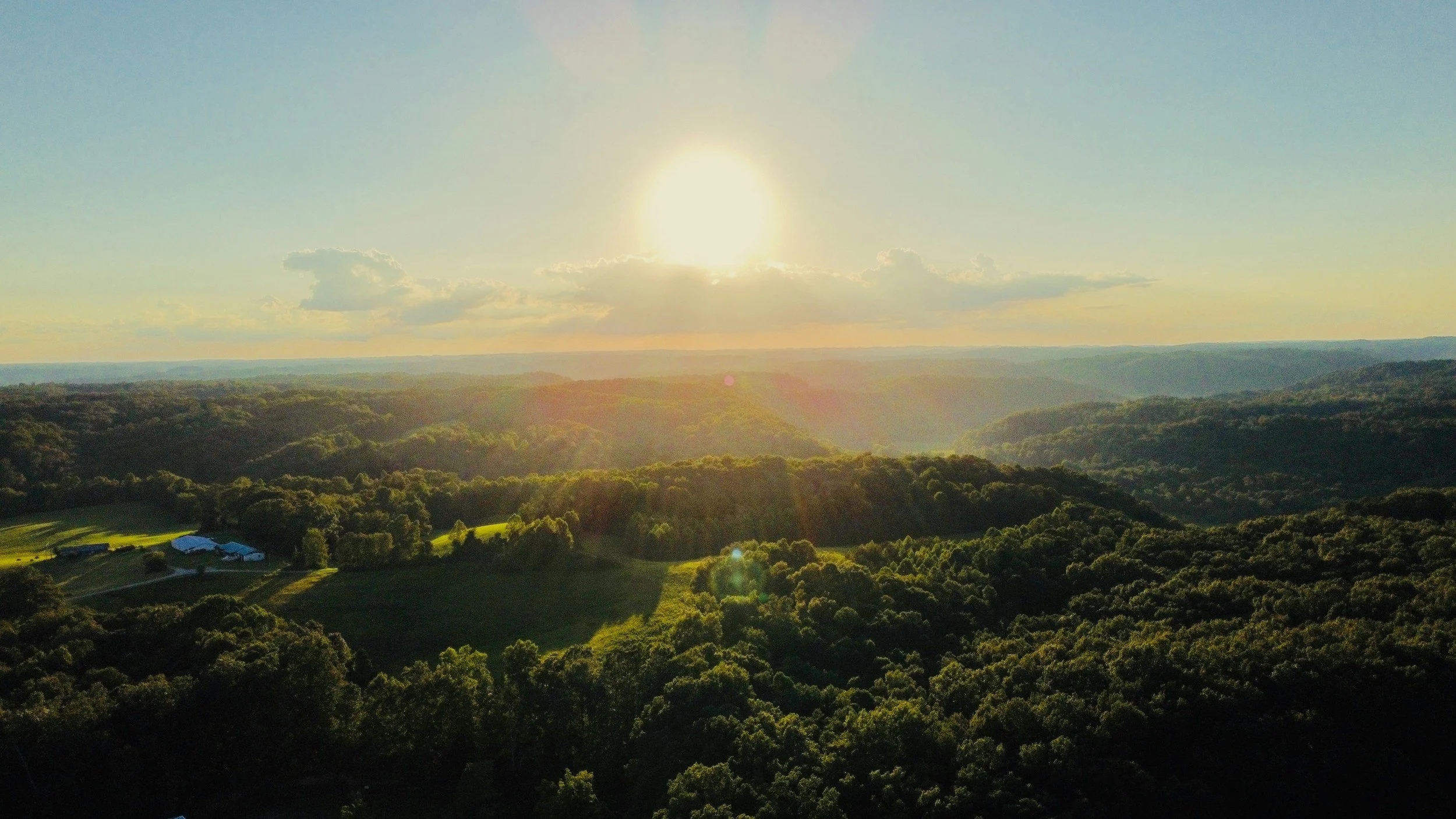 Landscape of rolling green hills and forests with a few buildings on the left, under a setting sun in a clear, partly cloudy sky.