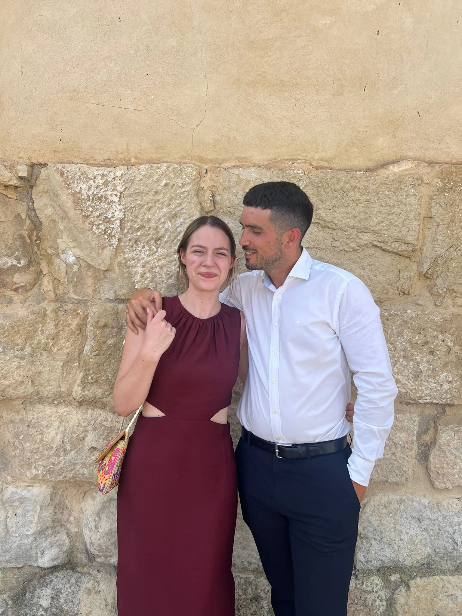 A man and woman standing close together against a stone wall, with the man embracing the woman. The woman is smiling slightly and the man is looking at her. She is wearing a burgundy dress with cutout details, and he is wearing a white dress shirt and dark pants.