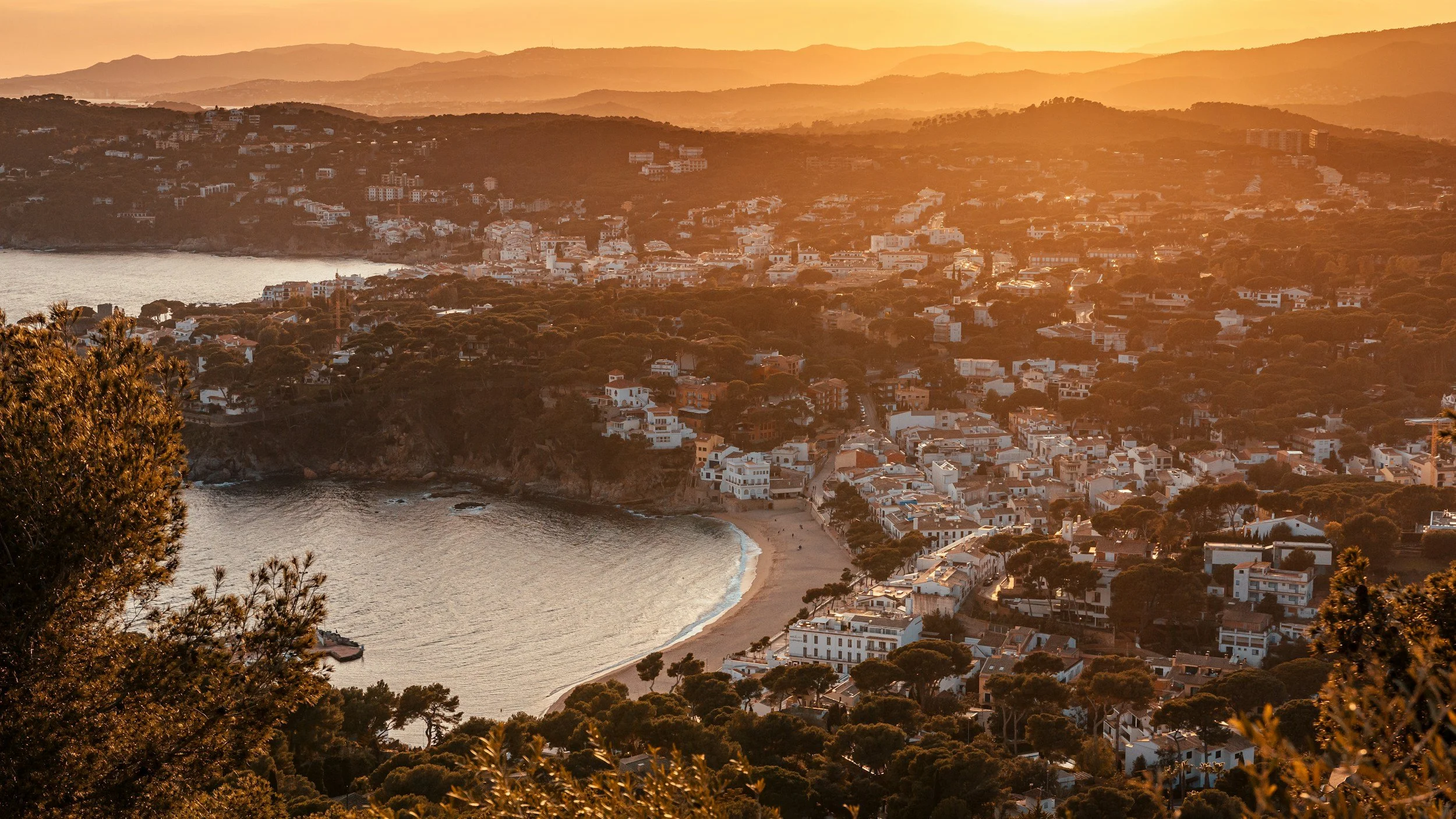 A coastal town at sunset with a crescent-shaped beach, surrounded by residential buildings and lush trees, with rolling hills in the background.