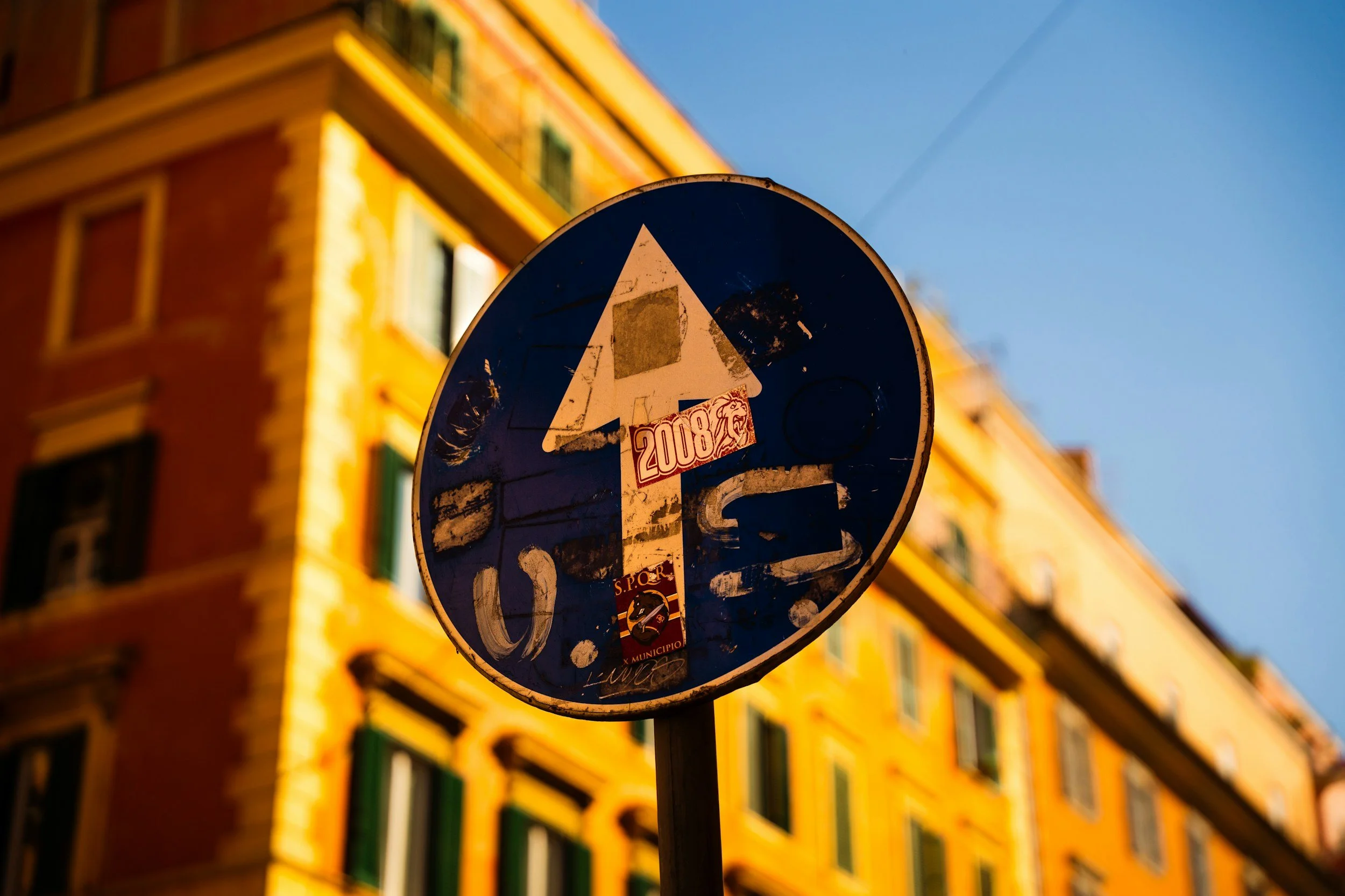 A weathered blue traffic sign with multiple decals and stickers, mounted on a pole in front of a colorful building with yellow walls and green window shutters, under a clear blue sky.