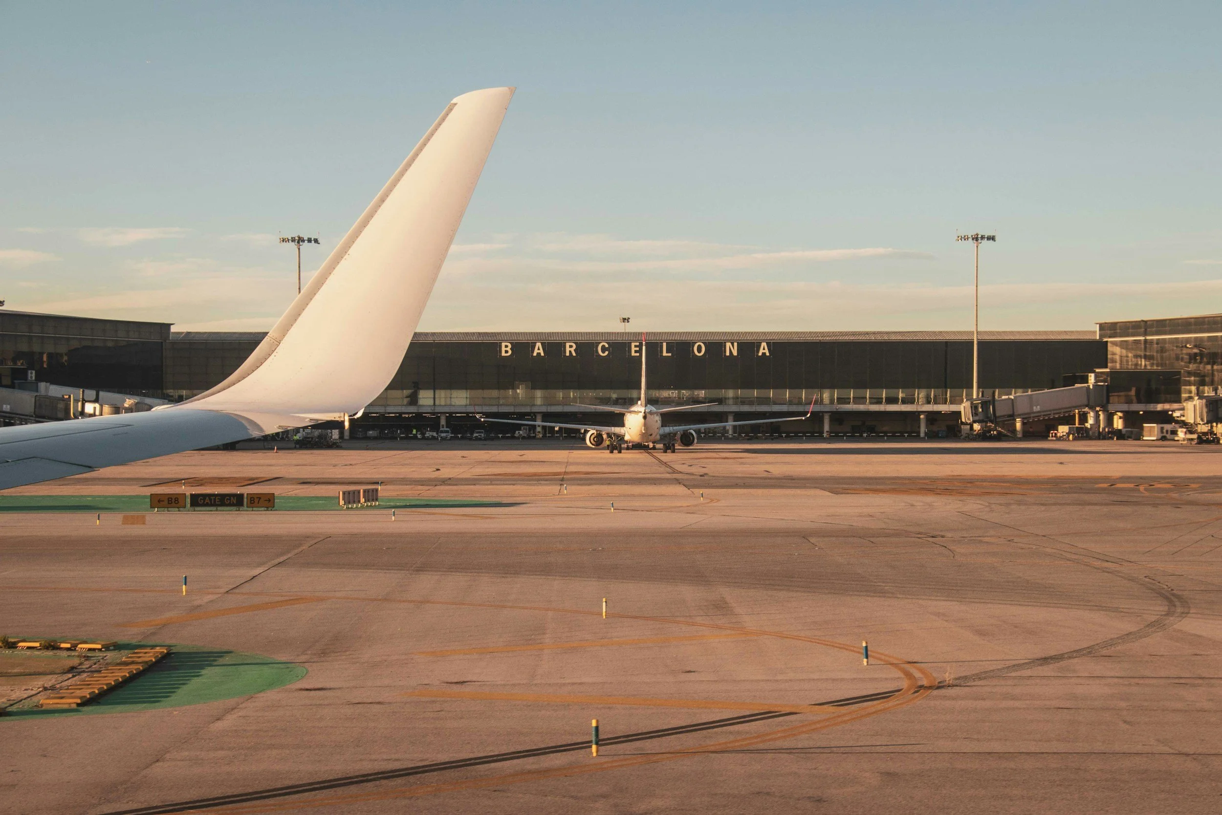 View of an airport tarmac with a tail of an airplane in the foreground, another airplane parked at the gate in the background, and a building with large windows displaying the word 'Barcelona'.