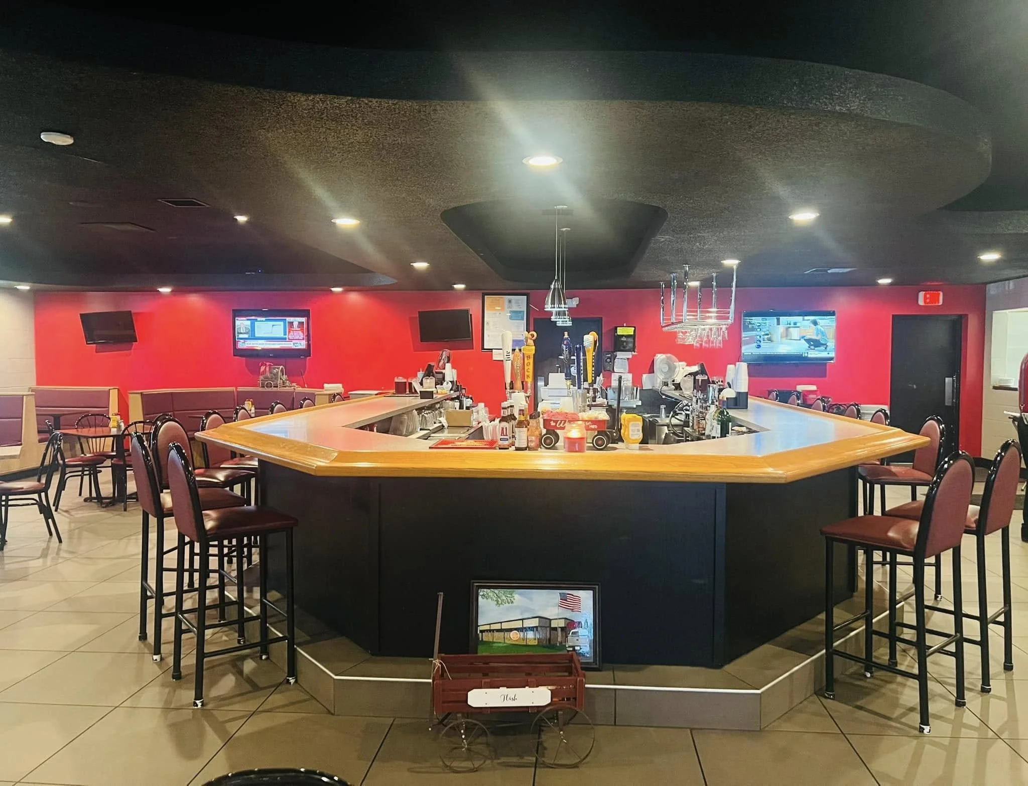 Inside a bar with a U-shaped counter, bar stools with red cushions, multiple television screens on a red wall, and drink-making equipment and bottles on the counter. Seating area with tables and chairs visible in the background.