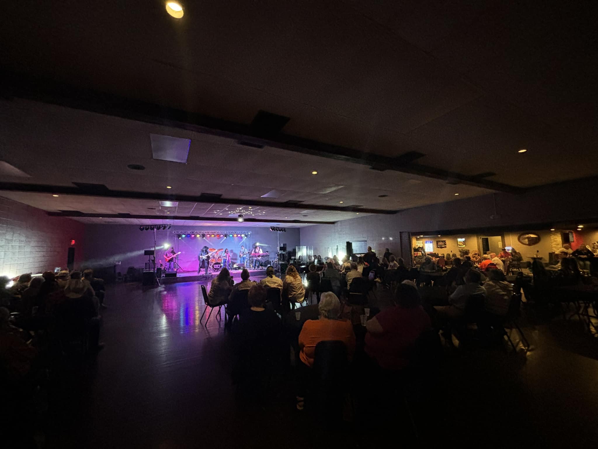 A live band performing on stage at an indoor venue with an audience seated in front of them. The stage is lit with colorful lights, and the venue has a dark, cozy atmosphere with some area lighting and a visible bar area on the right.
