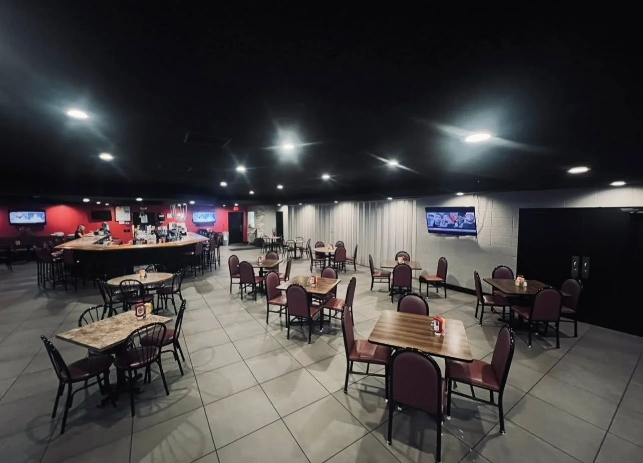 Empty restaurant seating area with tables and chairs, some tables with condiment bottles, television screens on walls, and a bar area in the background.