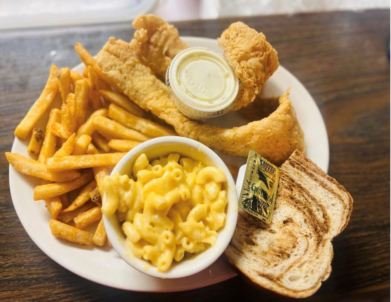 A plate of fried fish, French fries, macaroni and cheese, a slice of toasted bread, and a small container of tartar sauce.