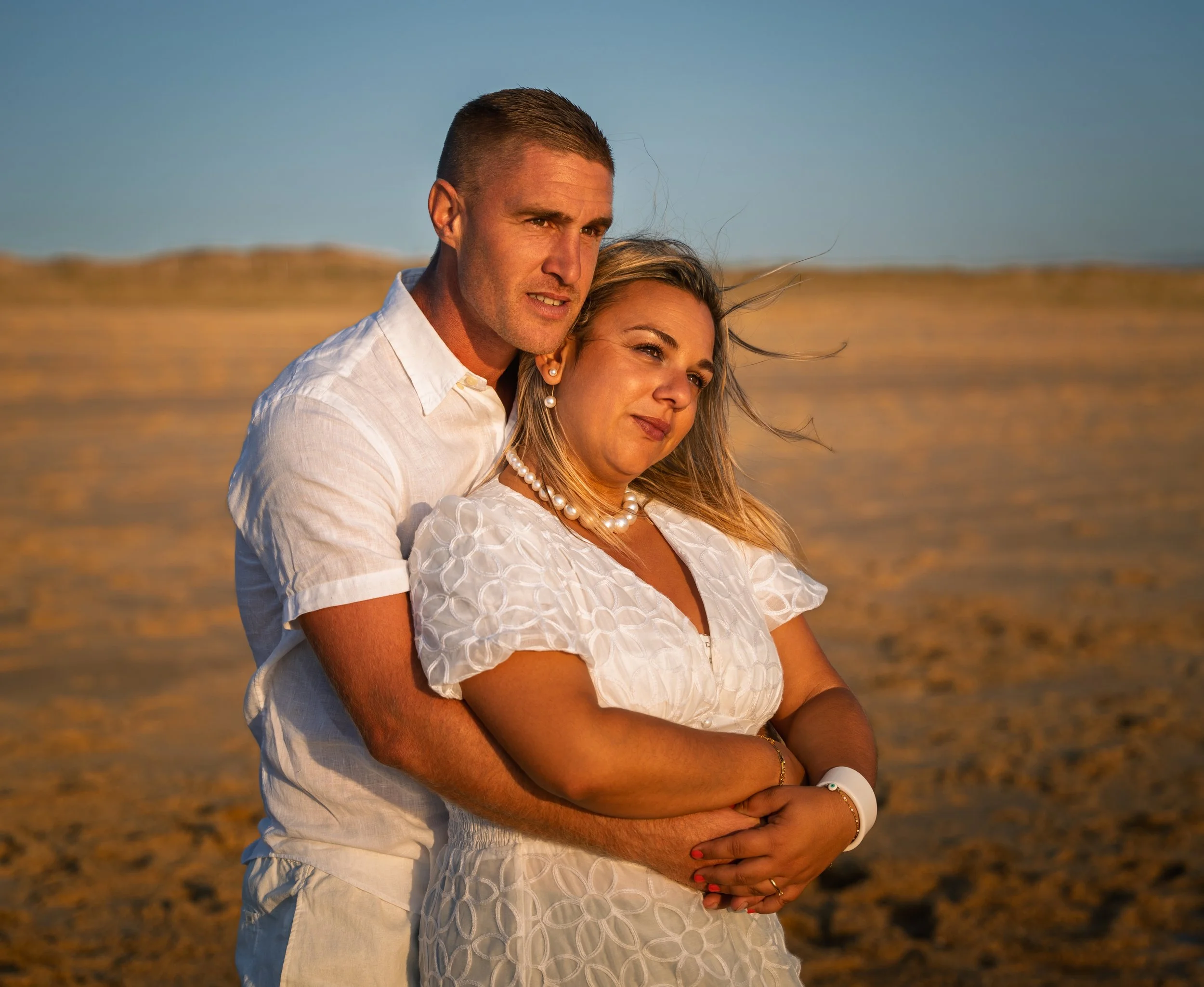 A couple standing on a beach during sunset, with the man embracing the woman from behind. Both are dressed in white, and the woman is wearing a pearl necklace and bracelet. The background shows sandy dunes and a clear blue sky.