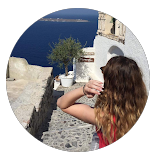 A woman with long brown hair standing on a cobblestone street, facing a white building with a ceramic pot on the wall, overlooking blue water.