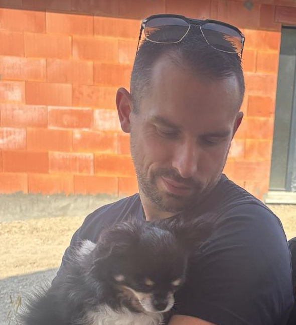A man with glasses on his head holding a small black and white dog outside near a brick wall.