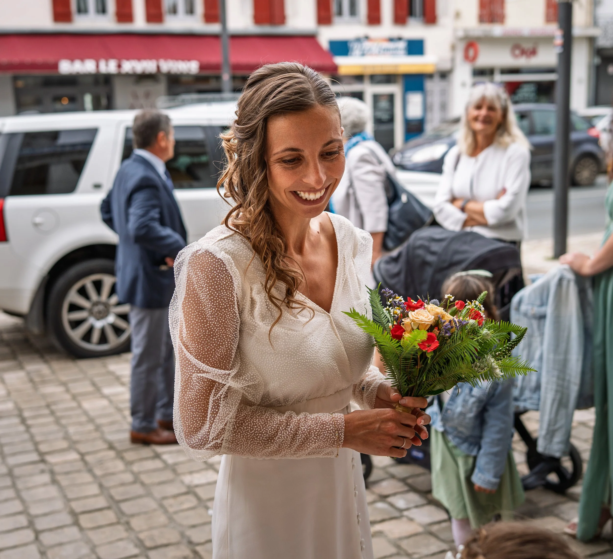A woman in a white dress holding a colorful bouquet of flowers, smiling, standing on a cobblestone street with people and cars in the background.