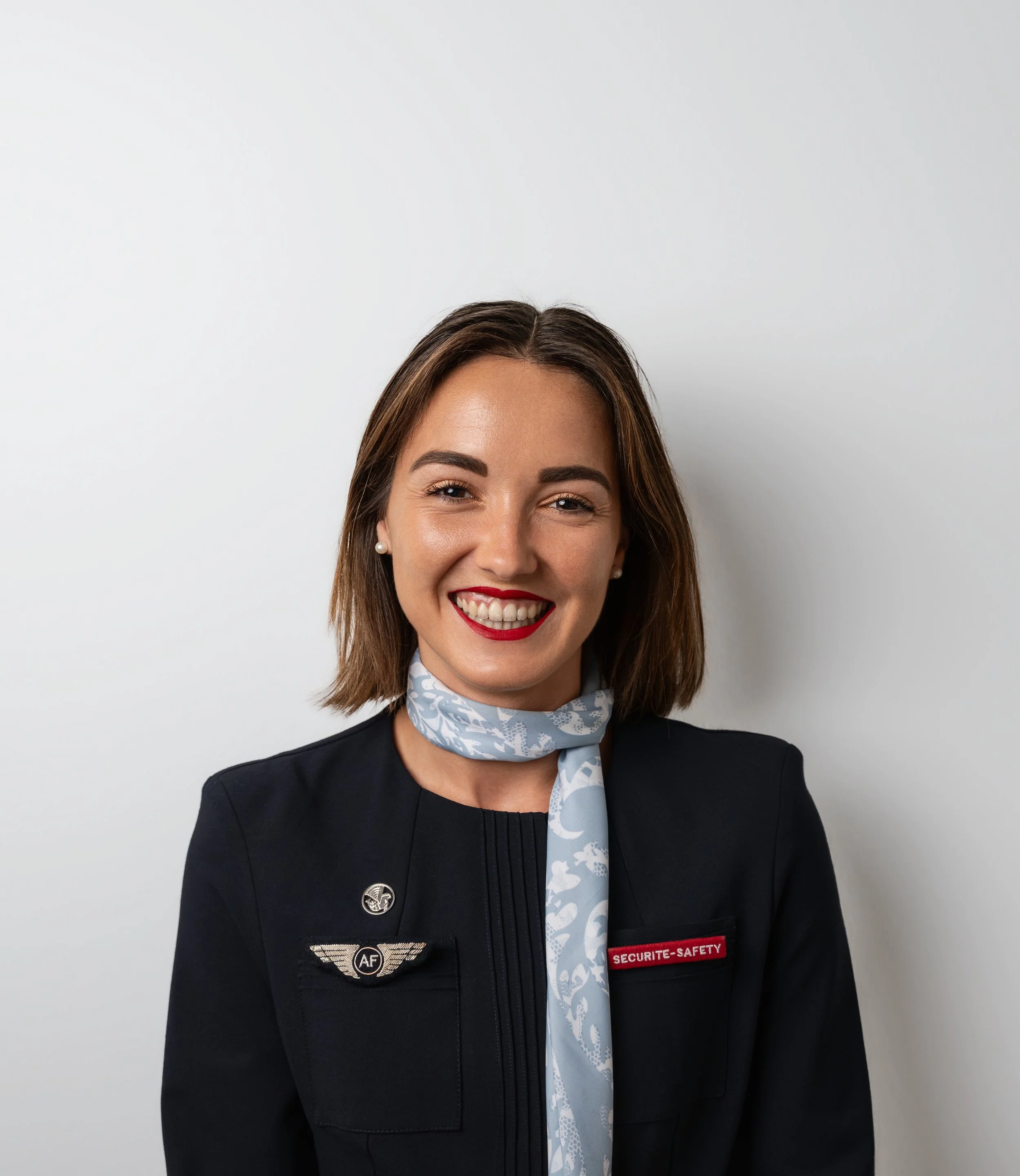 A smiling woman wearing a black uniform jacket with a wing pin and a name tag that says 'Securite-Safety', a patterned light blue scarf, and pearl earrings, standing against a white background.