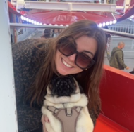 A woman with long hair and sunglasses smiling while holding a small dog in a harness at an amusement park ride.