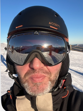 Close-up of a person wearing a black helmet with the brand 'Aclaimed' and ski goggles in a snowy mountain landscape during sunset.